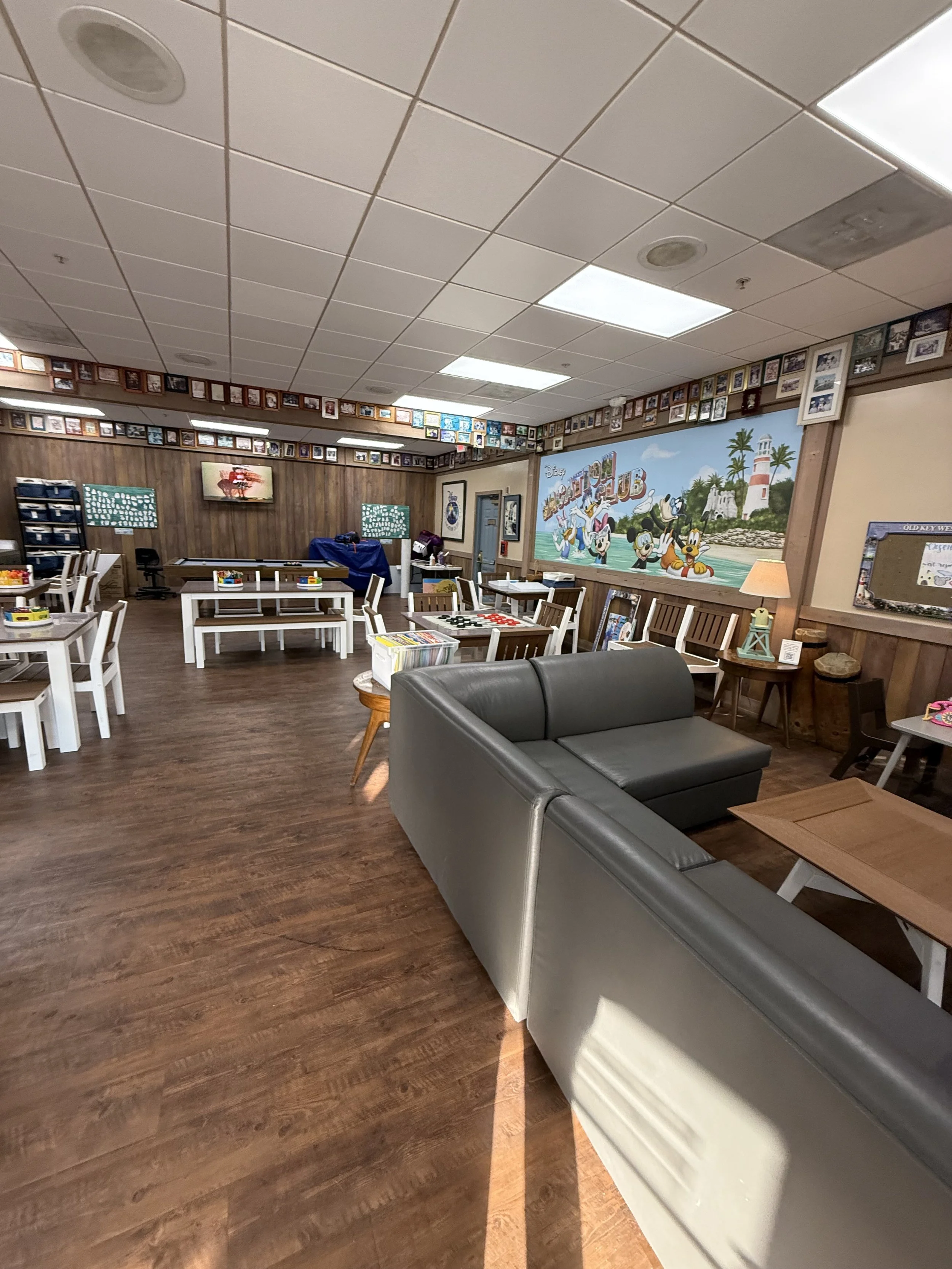 Empty children and family gathering room with wooden flooring, white and black furniture, Disney themed mural, and numerous framed pictures on the walls.