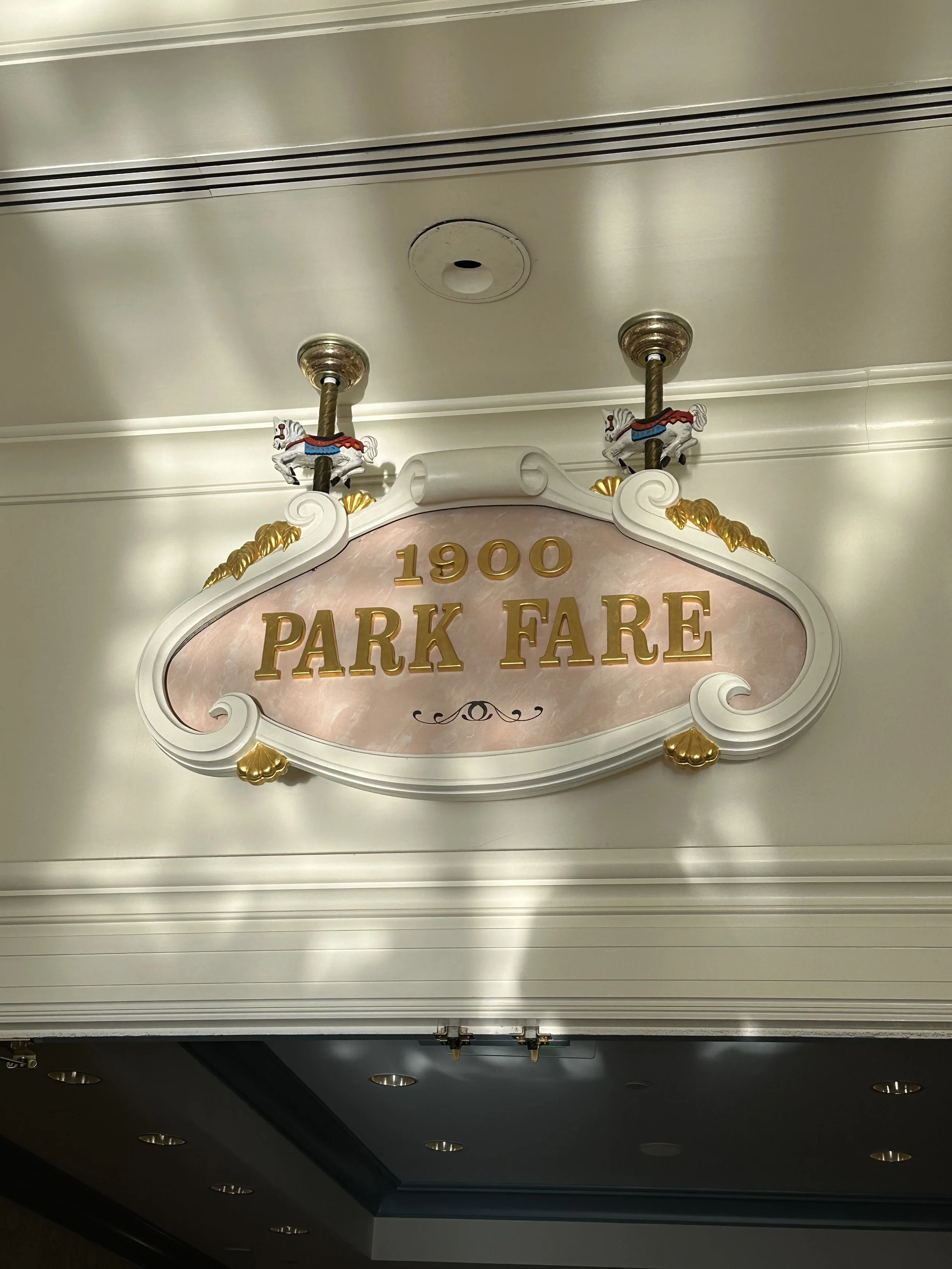 Vintage sign reading '1900 PARK FARE' with gold lettering, decorative gold accents, and carousel horse ceiling mounts.
