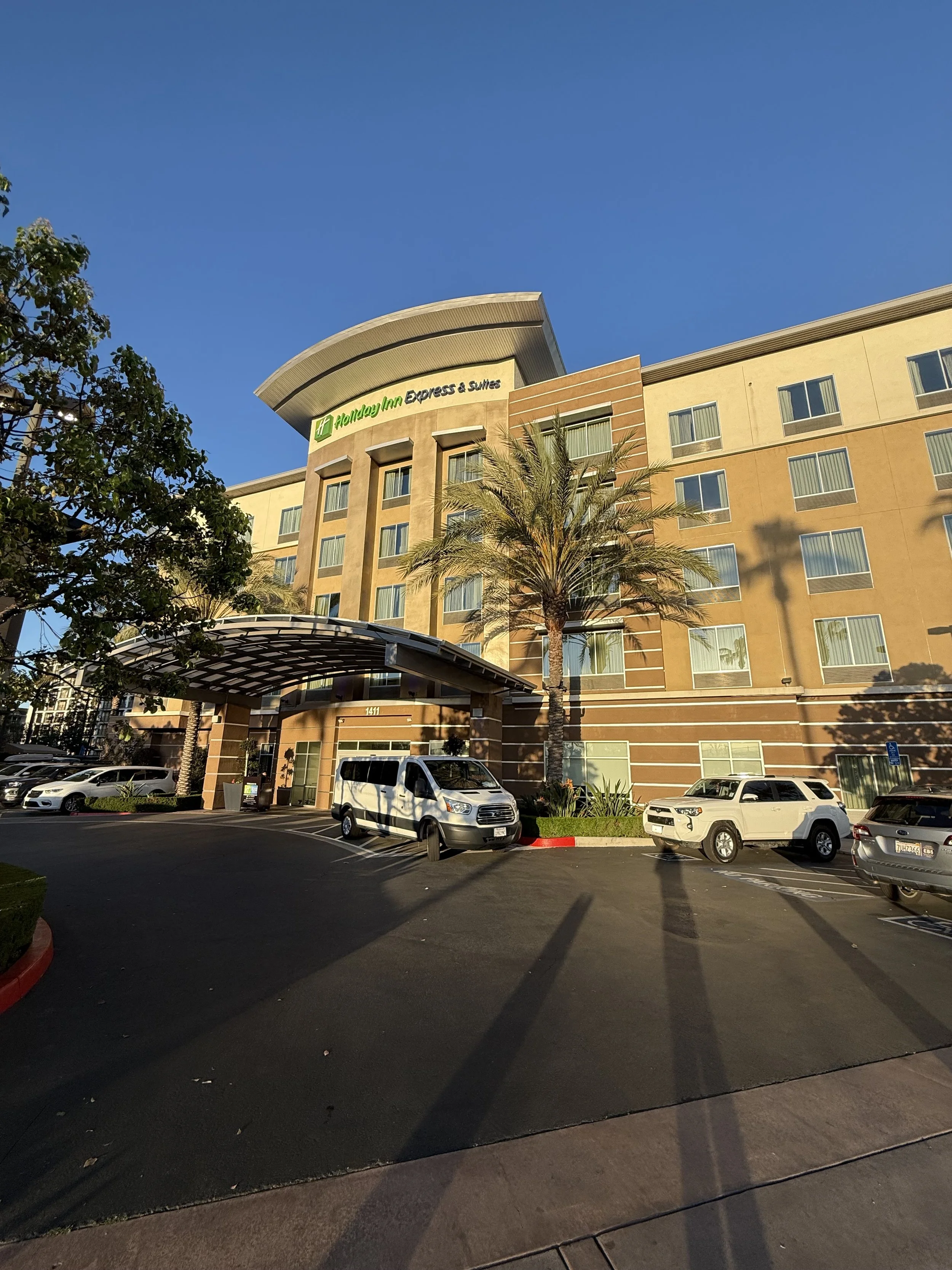 Front of a Holiday Inn Express & Suites hotel with parked cars and palm trees under a blue sky.