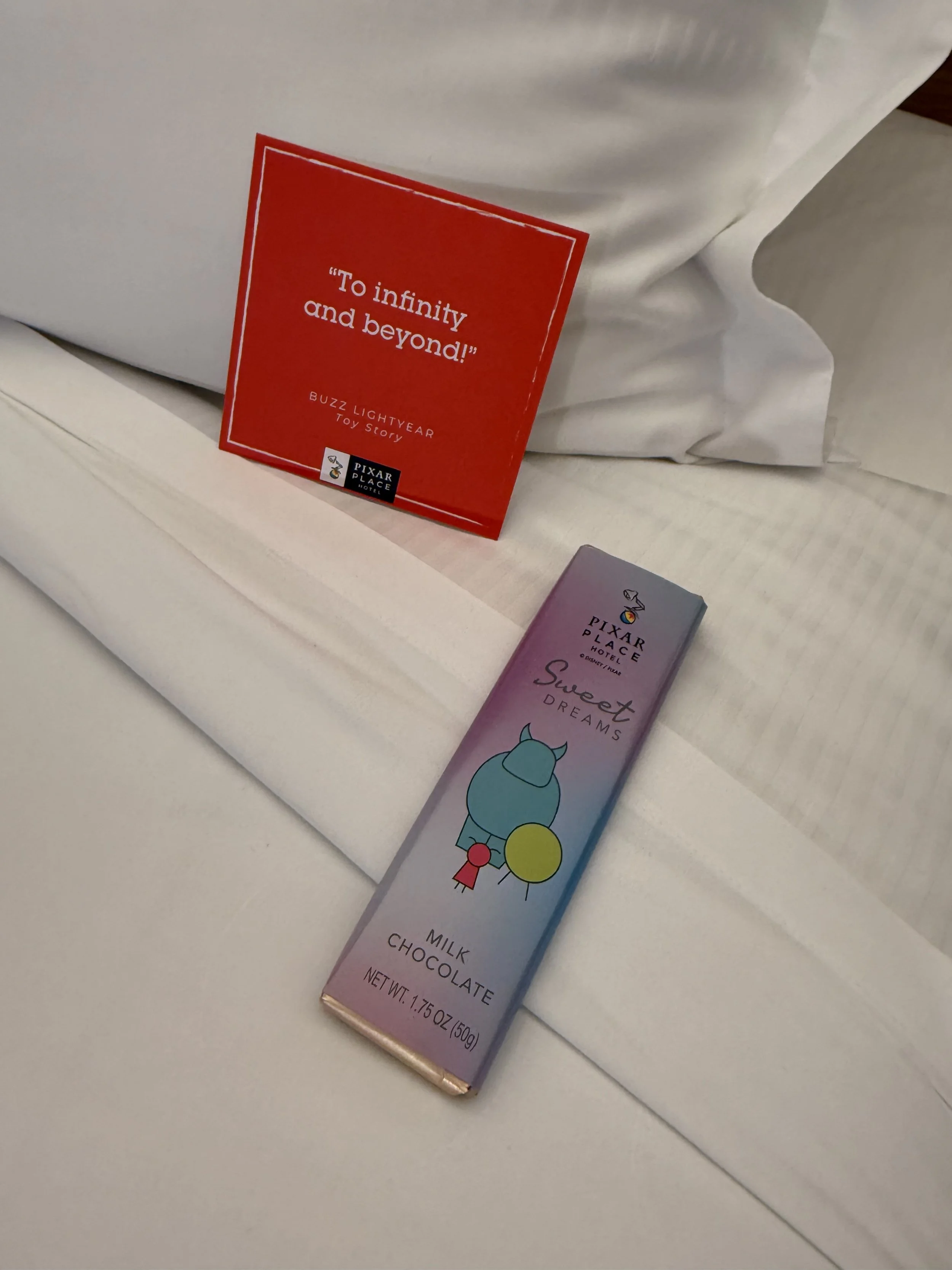 A pink and purple box of Sweet Dreams milk chocolate with a cartoon character on it, placed on a white bed beside a white pillow and a red informational card with white text.