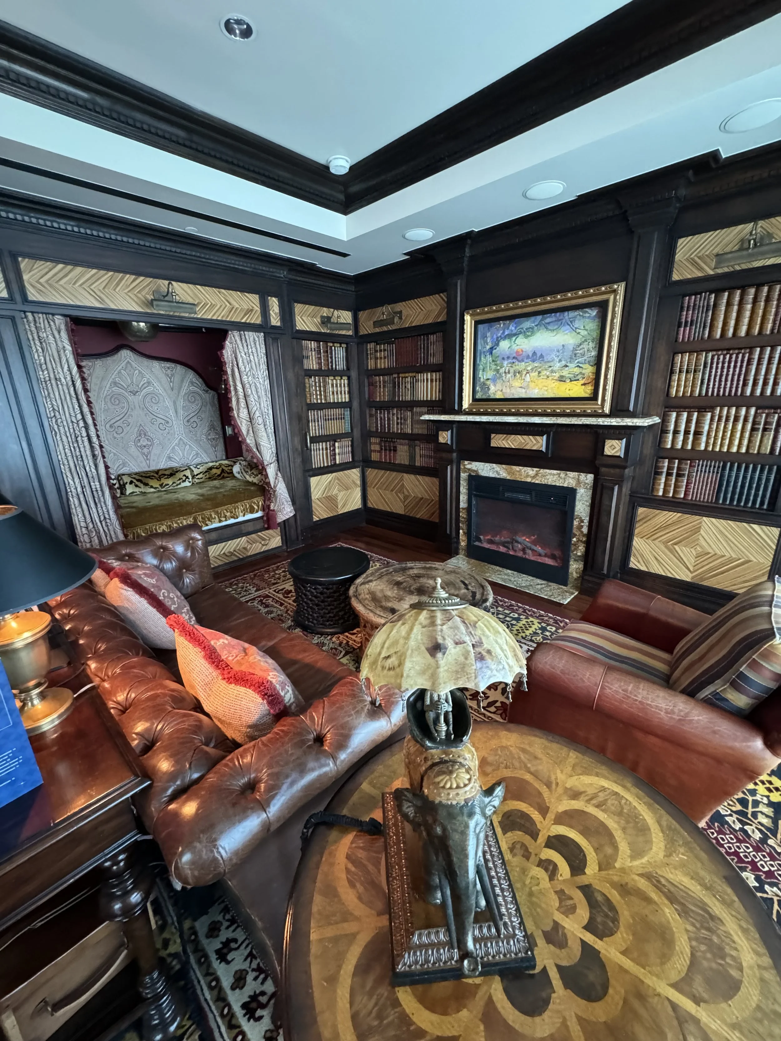 A cozy living room featuring leather sofas, a patterned wooden table with a decorative sculpture and lamp, a fireplace with a framed landscape painting above, and built-in bookshelves filled with books. There's an alcove with a cushioned seat and cur