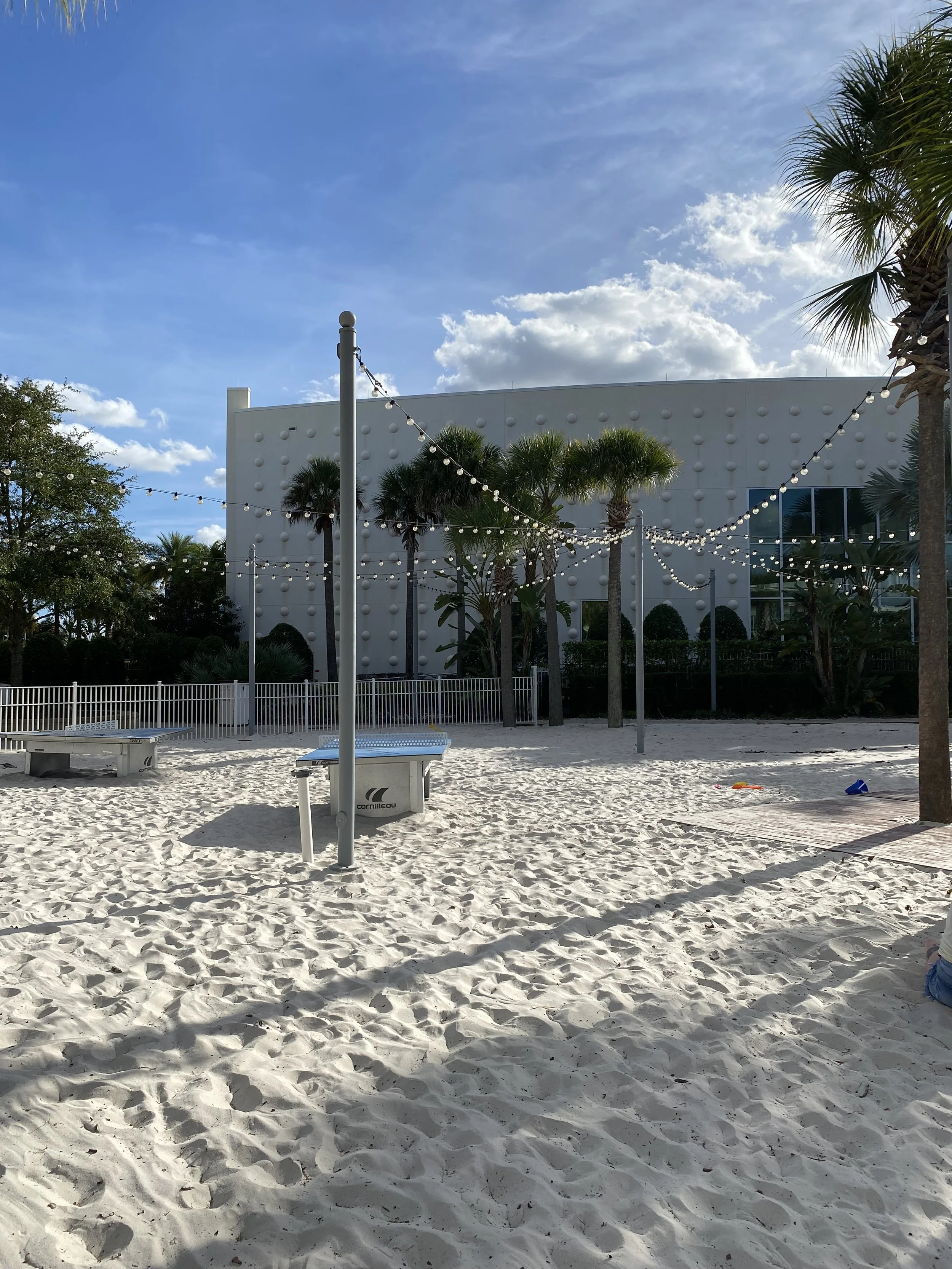 Empty beach volleyball court with sand, surrounded by palm trees and string lights, against a building with a modern white facade and a blue sky with clouds.