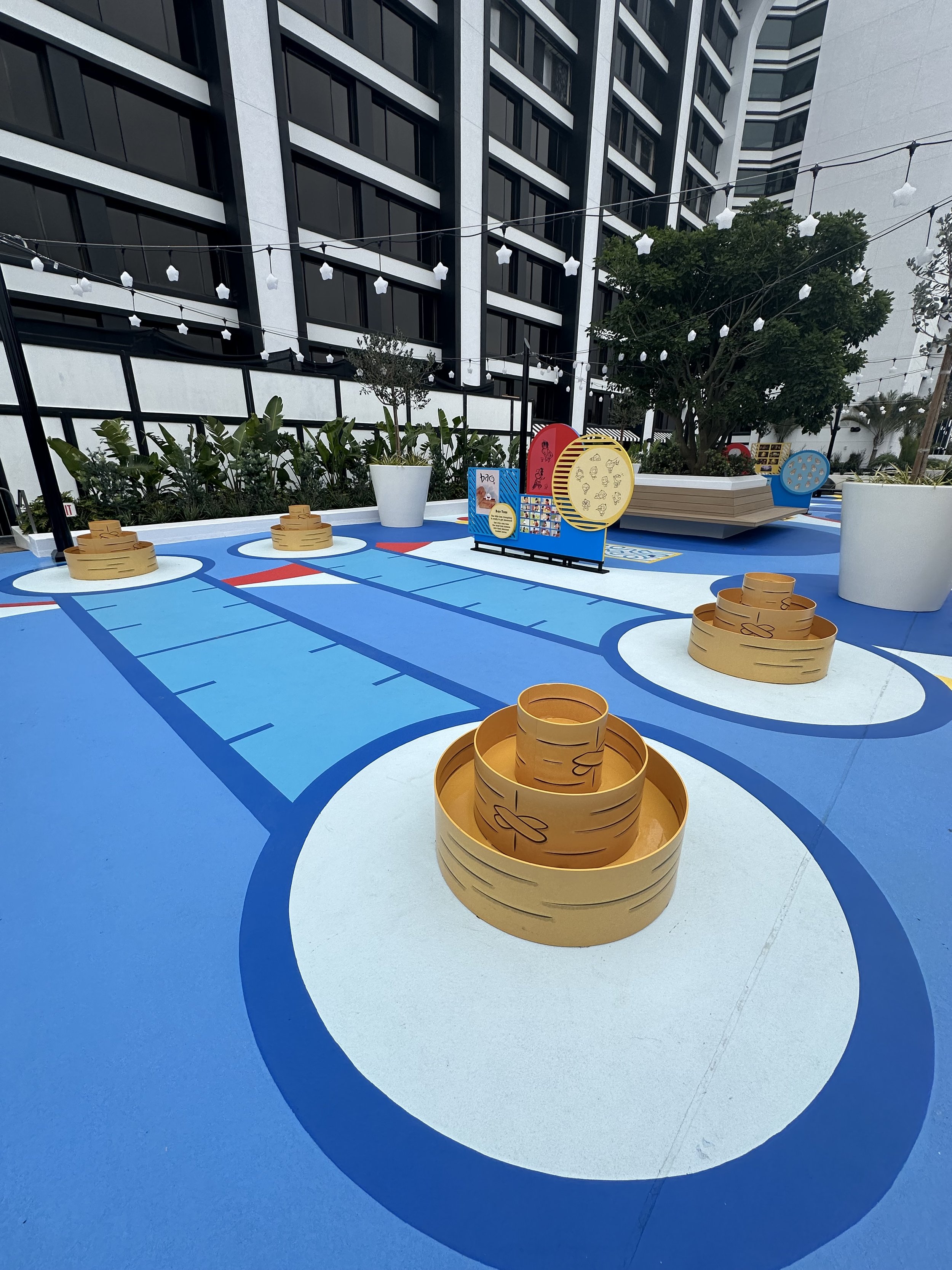 Outdoor mini-golf putting course with blue and white sections, decorative orange stacks, potted plants, and string lights, with a modern building in the background.