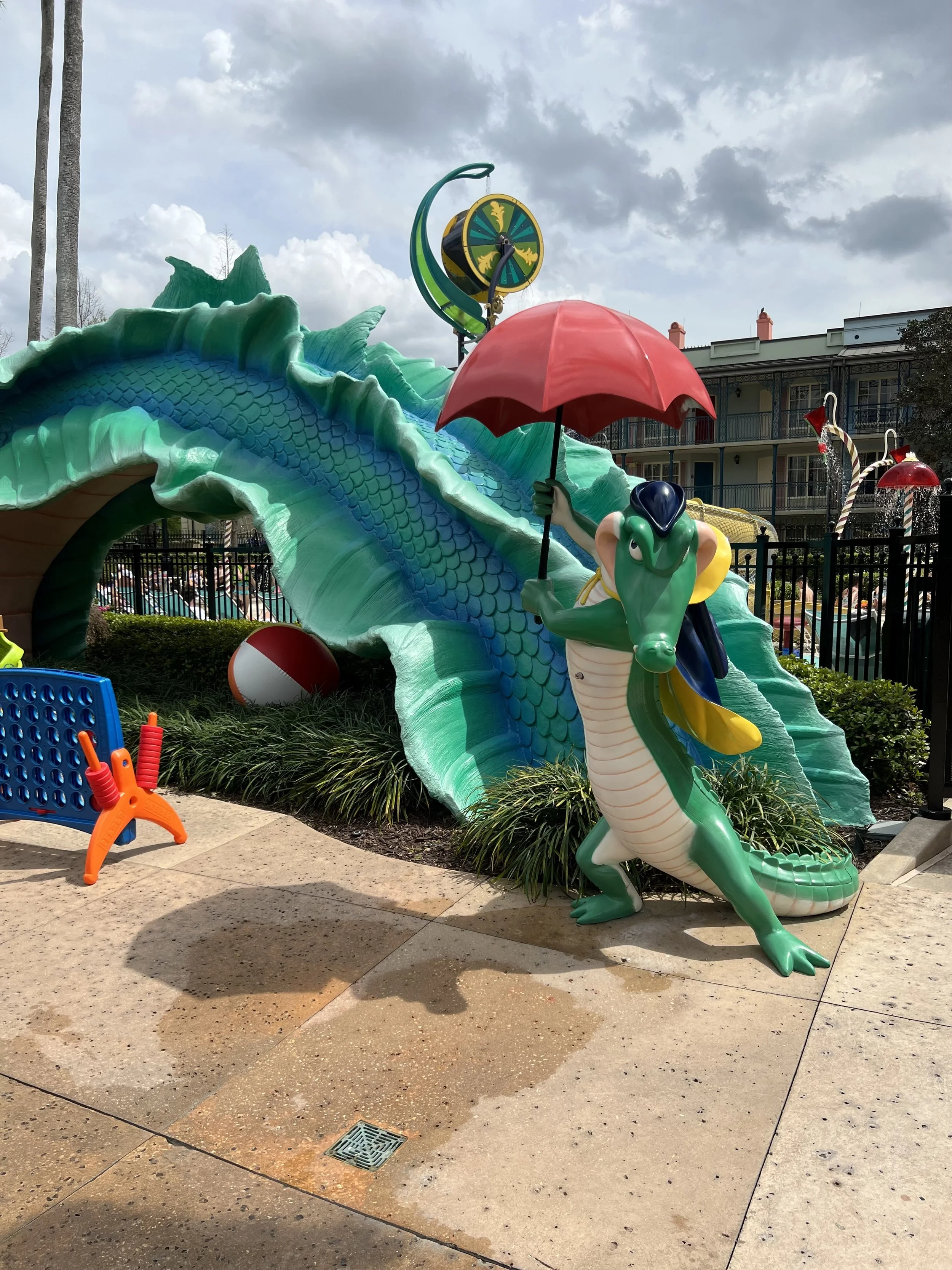 A whimsical sculpture of a green alligator holding a red umbrella, standing in front of a large, animated blue dragon with green accents. The scene includes a beach ball and a parking lot with buildings in the background.