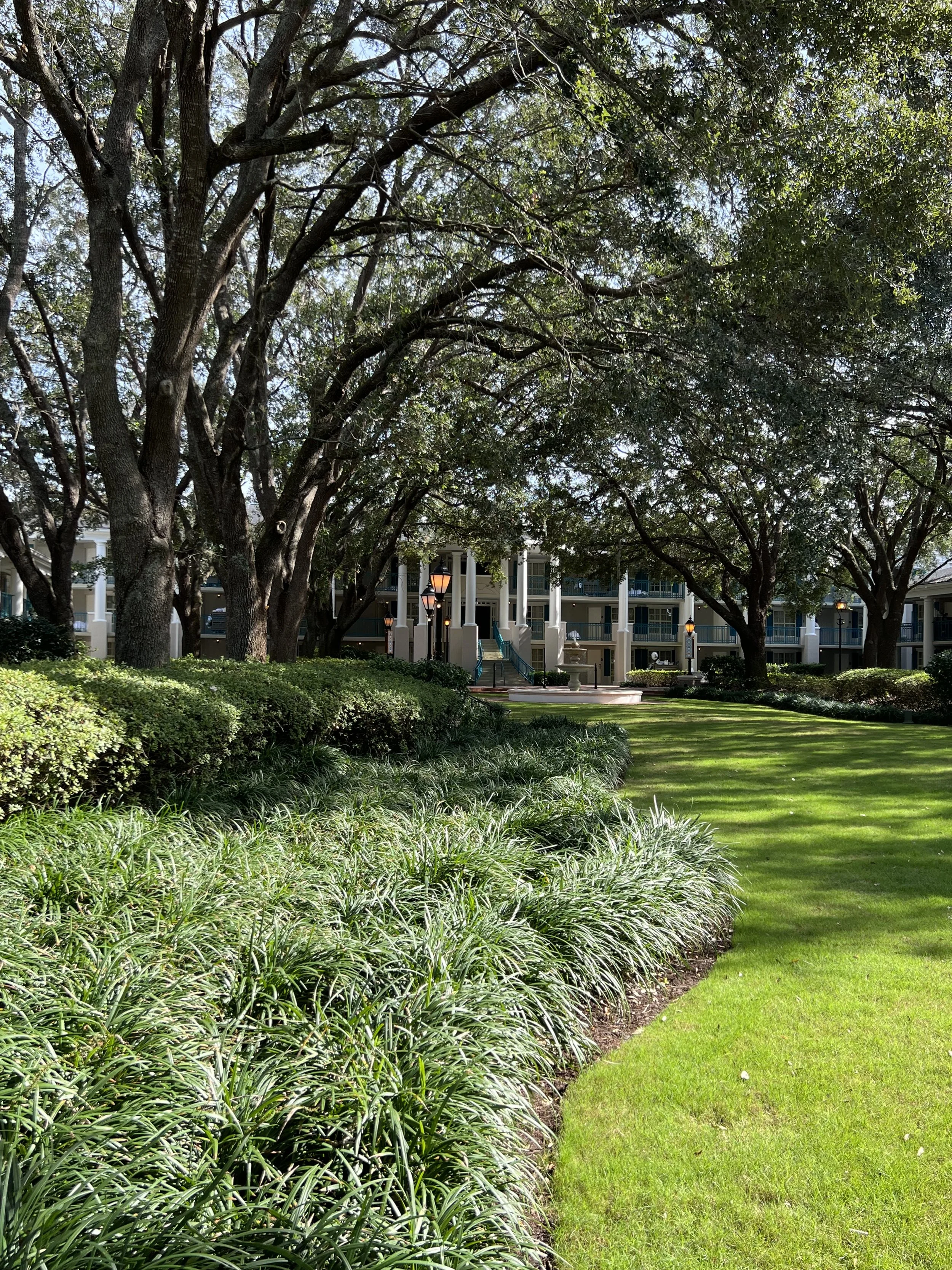 A lush manicured lawn with edging plants and bushes, tall trees providing shade, and a multi-story residential building with balconies and street lamps in the background.
