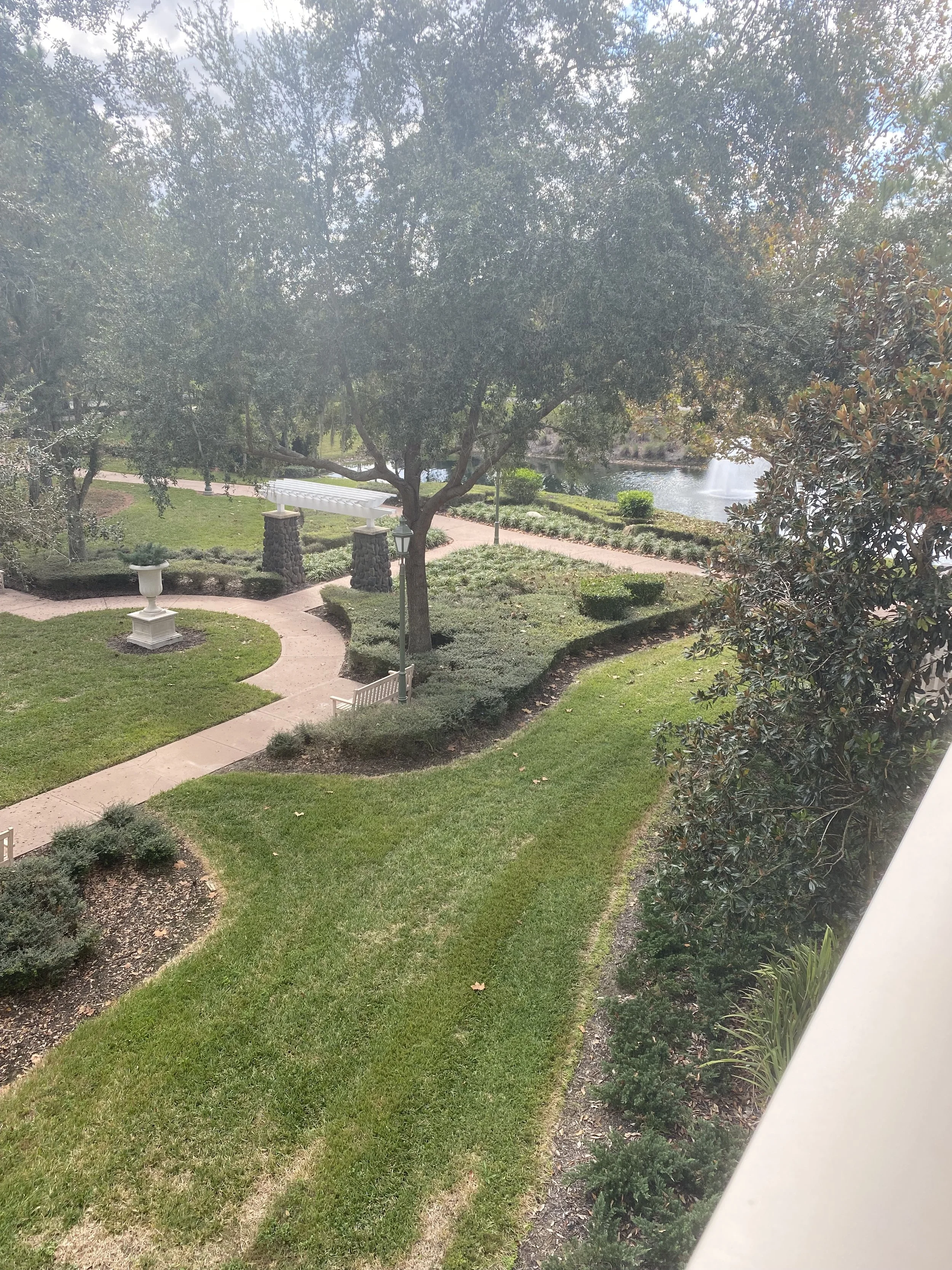 A well-maintained park with lush green grass, trees, a winding pathway, benches, a fountain in the background, and a body of water with a fountain.