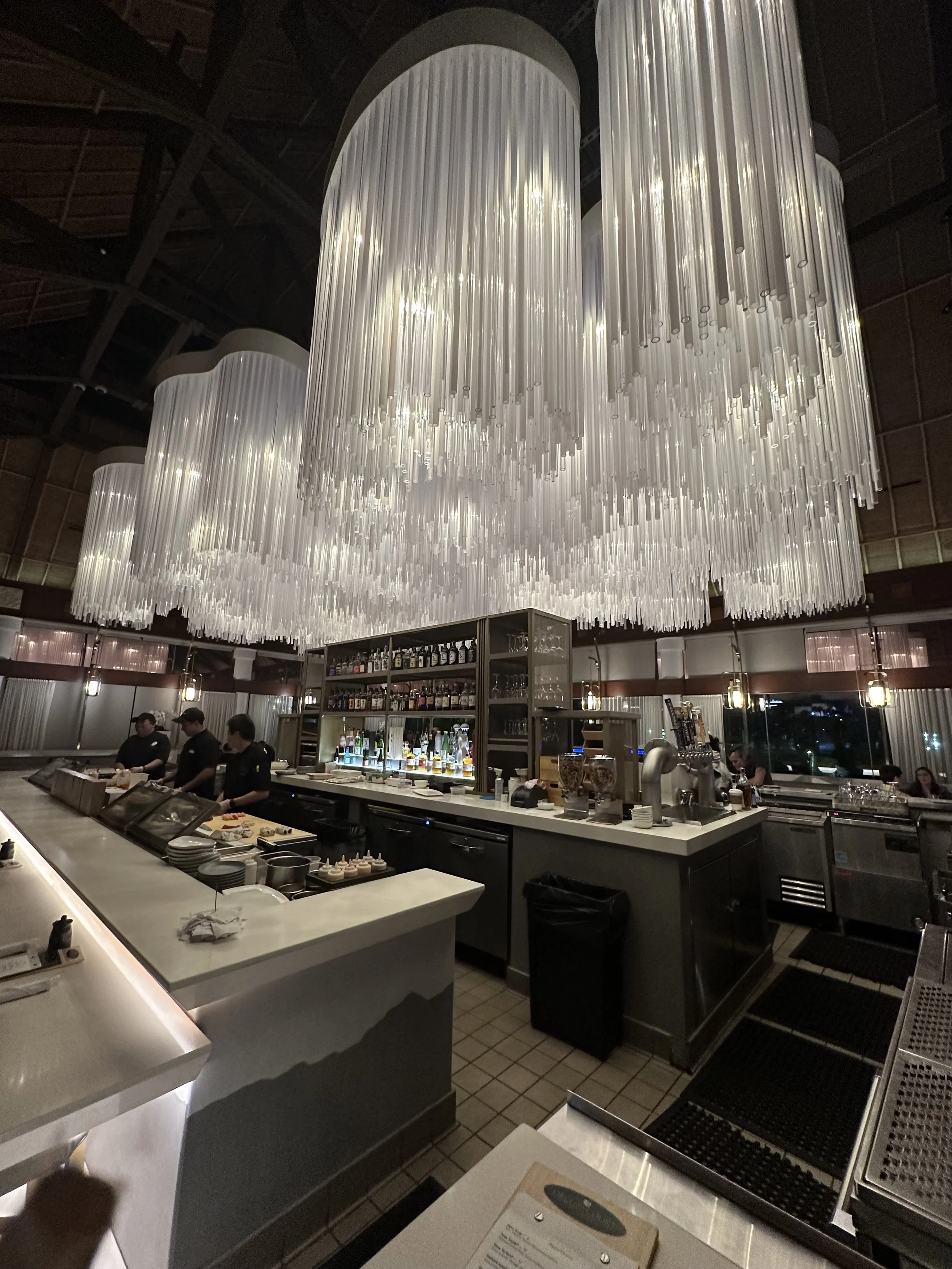 Elegant bar with large, modern, chandelier lighting fixtures hanging from the ceiling, bar counter with drinks, bar staff, and patrons seated at tables.