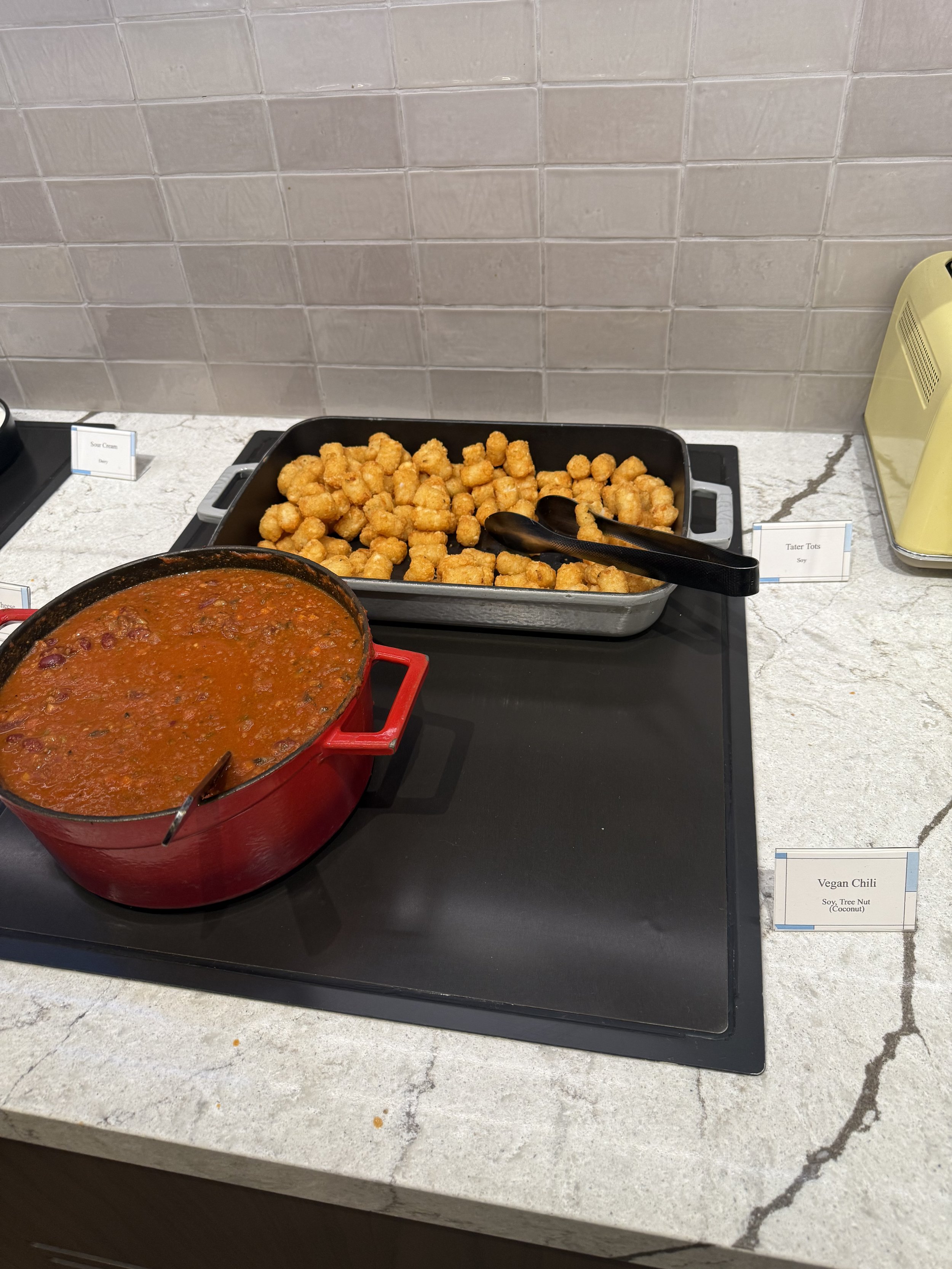 Buffet with a pot of vegan chili and a tray of tater tots on a marble countertop.