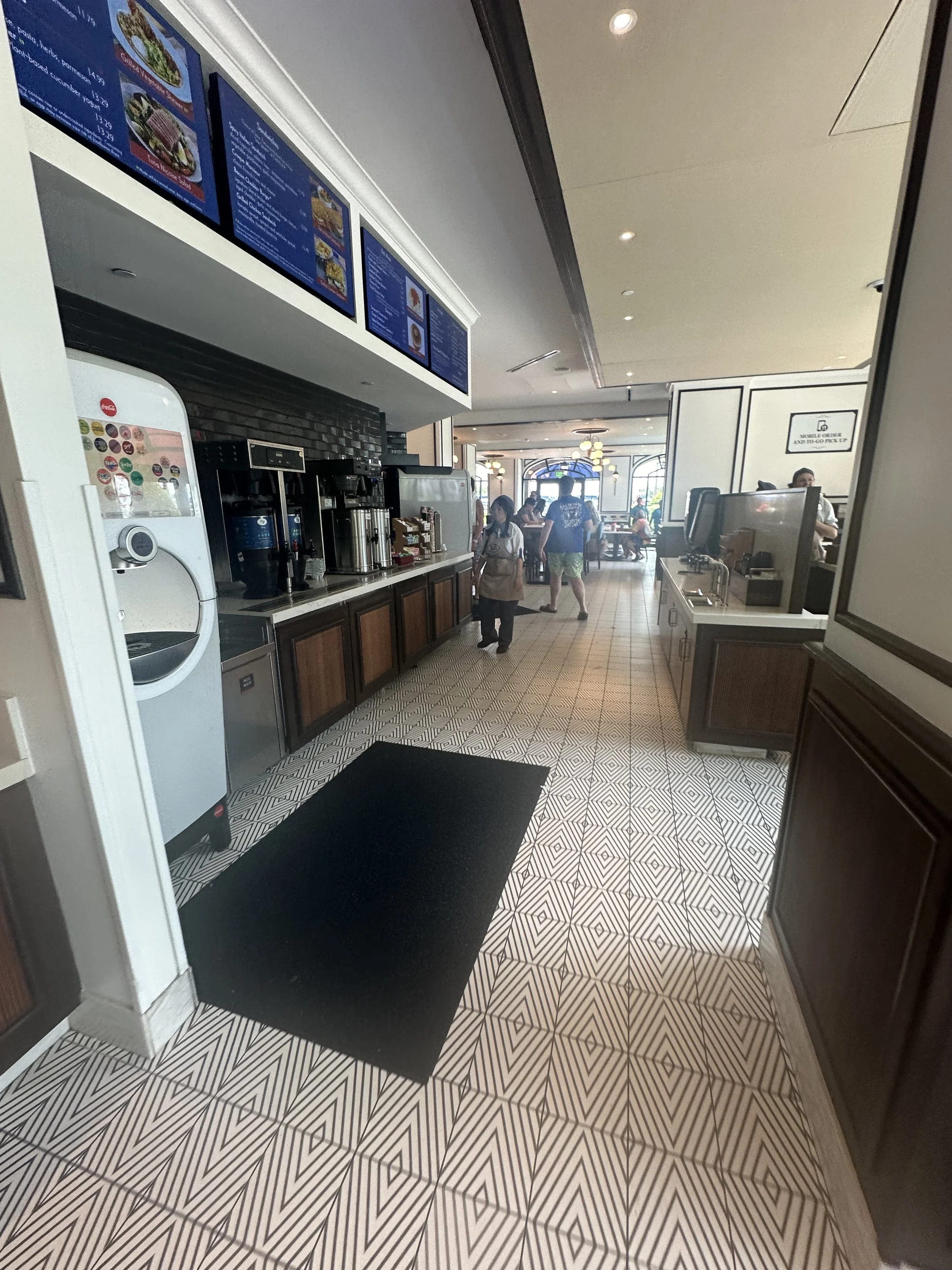 Inside a restaurant or cafe with a beverage station and menu screens, patterned tile floor, and customers in the background.