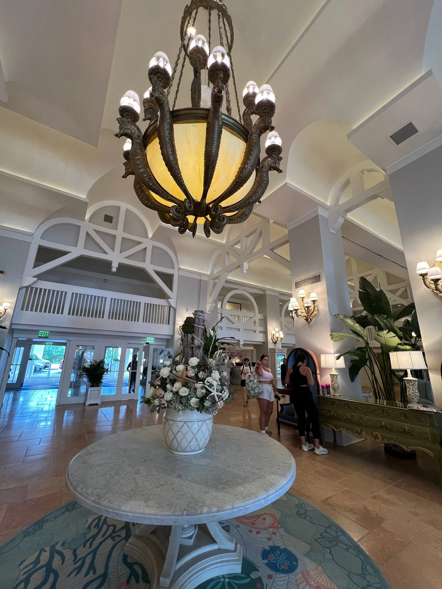 The image shows an elegant hotel lobby with a large ornate chandelier hanging from the ceiling, a round table with a floral arrangement in a ceramic vase in the foreground, and people standing near a reception desk in the background.