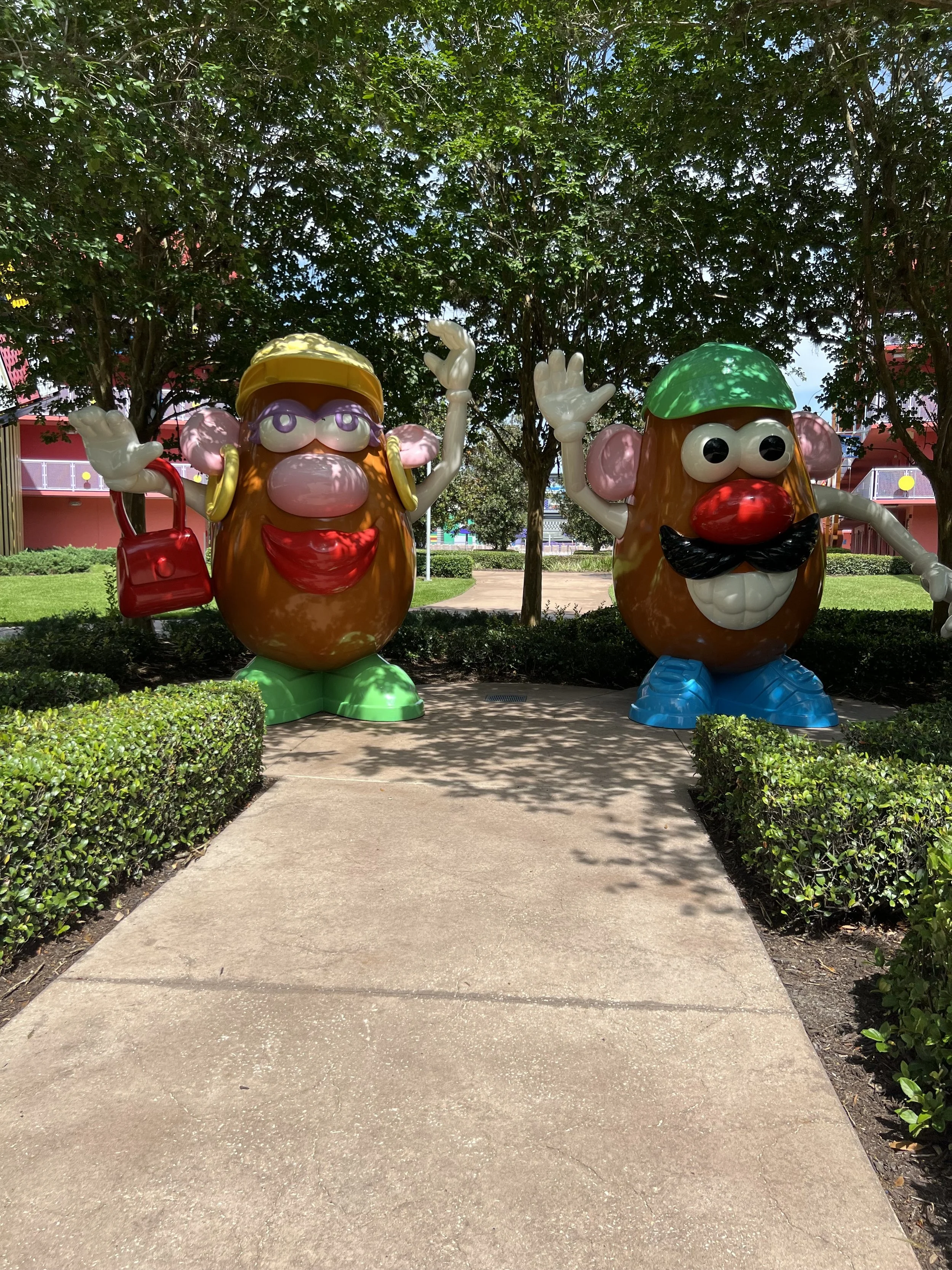 Colorful cartoon potato characters with faces, arms, and legs, standing outdoors under trees, with a sidewalk in foreground.