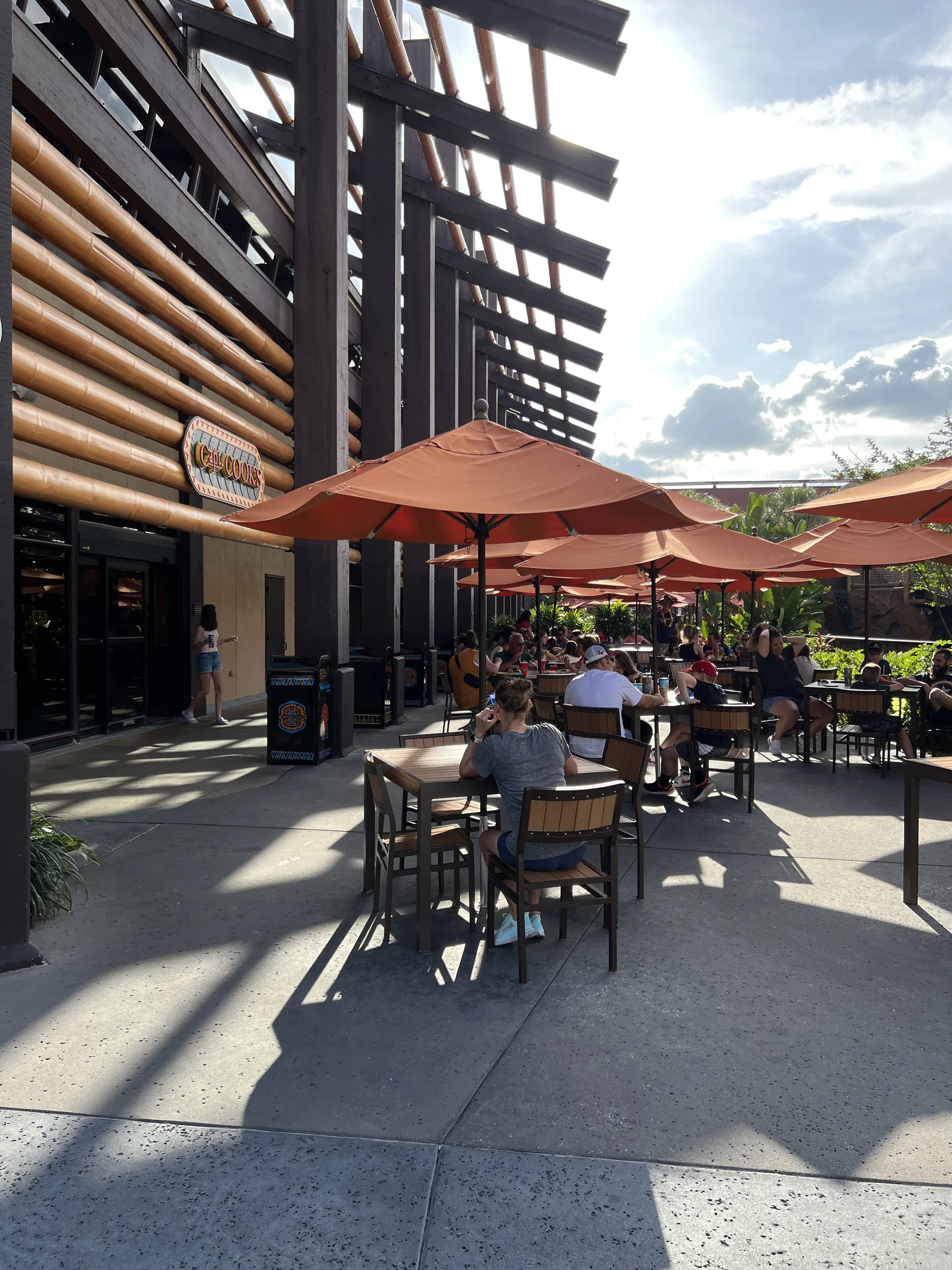 Outdoor patio at a restaurant with people sitting at tables under large orange umbrellas, some eating and talking, with a modern building in the background and cloudy sky overhead.
