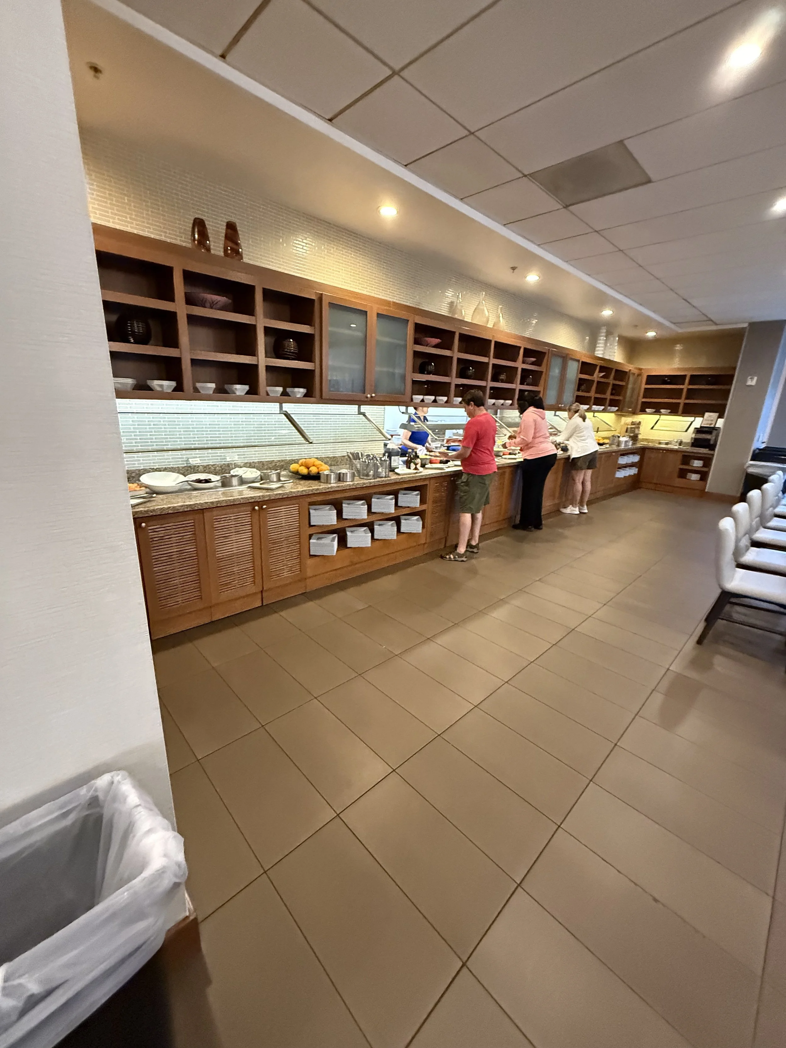 People serve themselves food at a buffet in a restaurant with wooden cabinetry and tiled flooring.