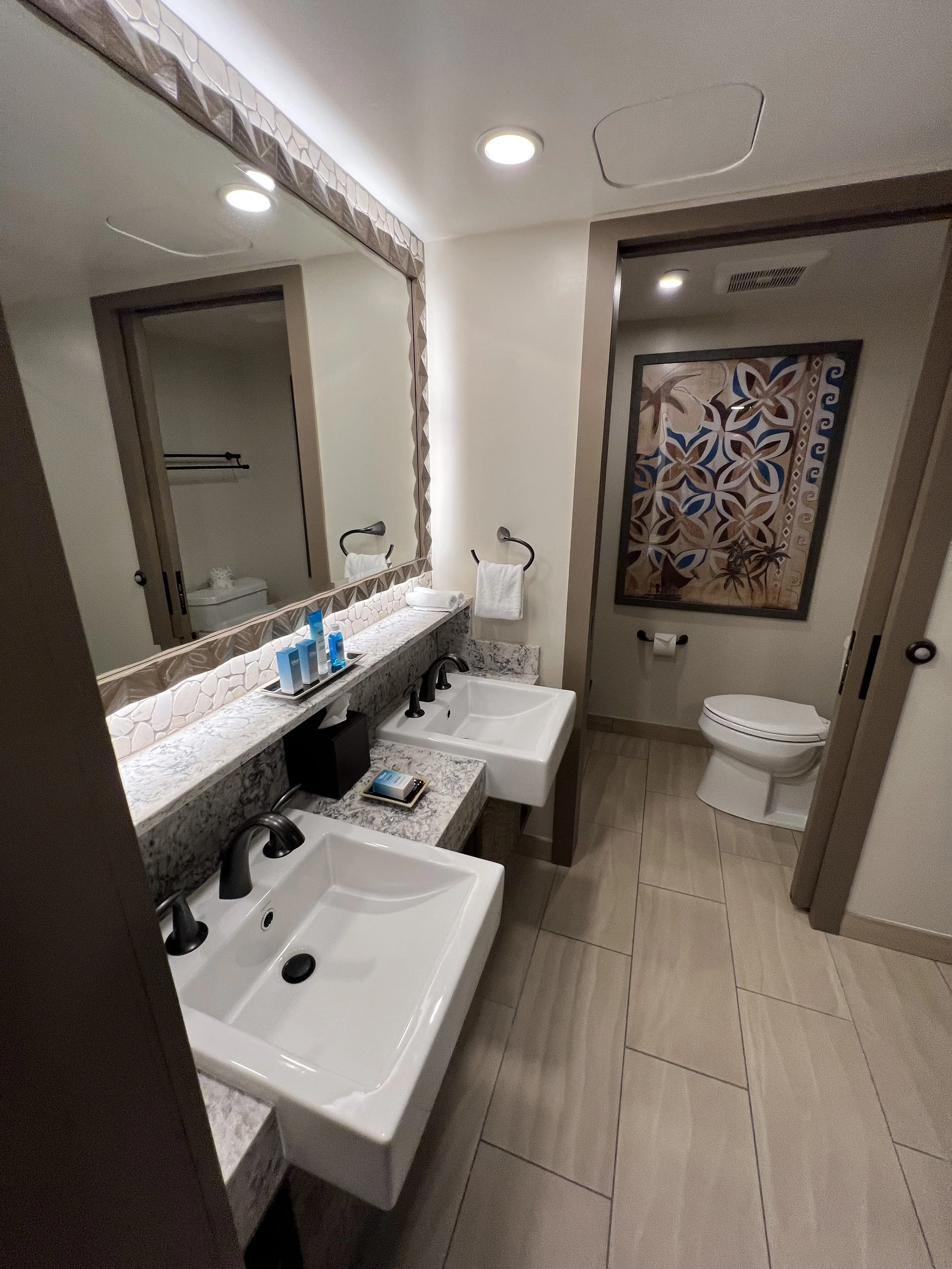 A bathroom with dual sinks, a large mirror, toiletries, and towels, with a separate toilet area featuring a piece of wall art.