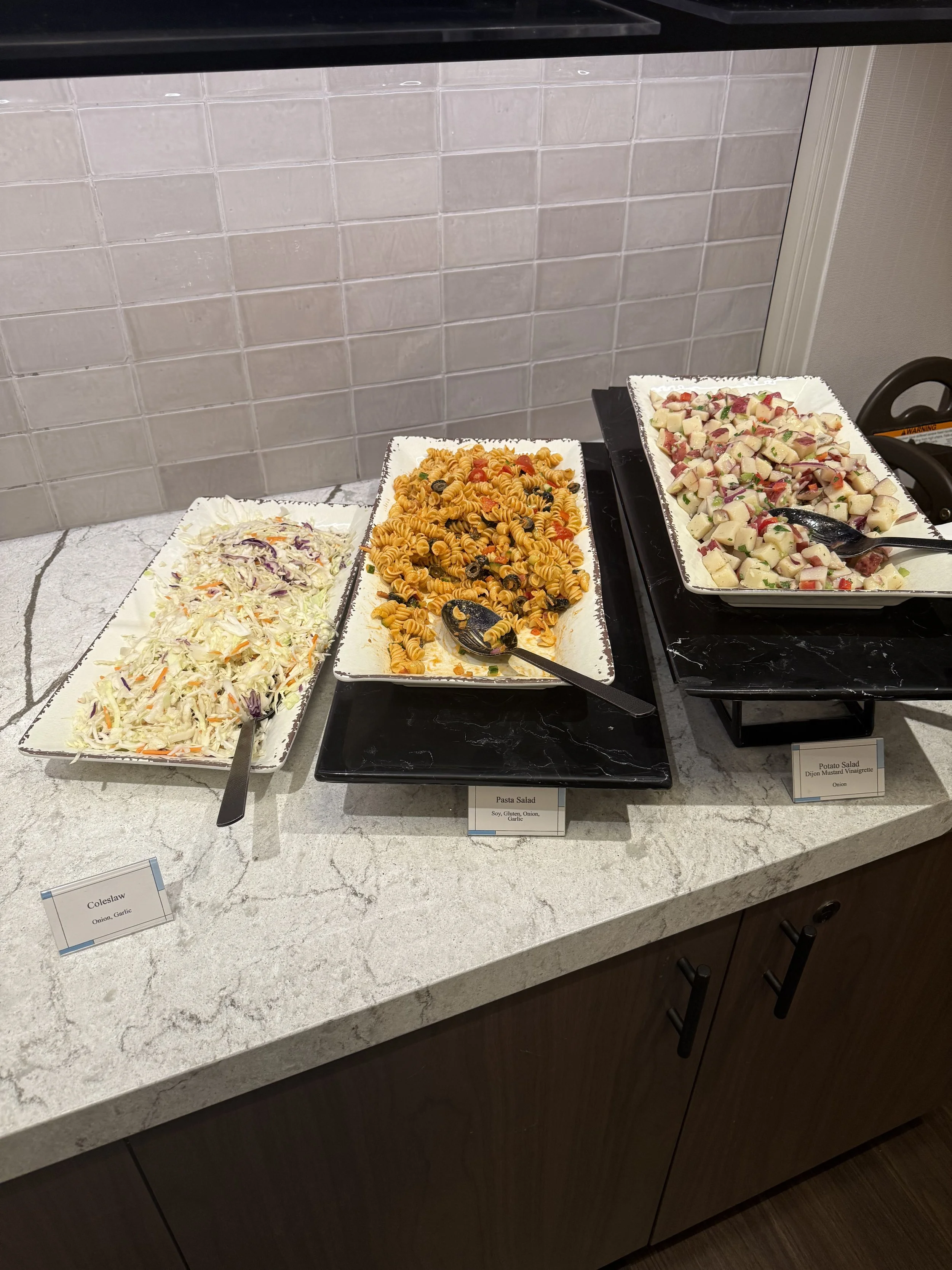 Three trays of salads on a marble counter. The left tray contains coleslaw, the middle tray has pasta salad with rotini pasta, and the right tray holds potato salad with diced potatoes, onions, and herbs.