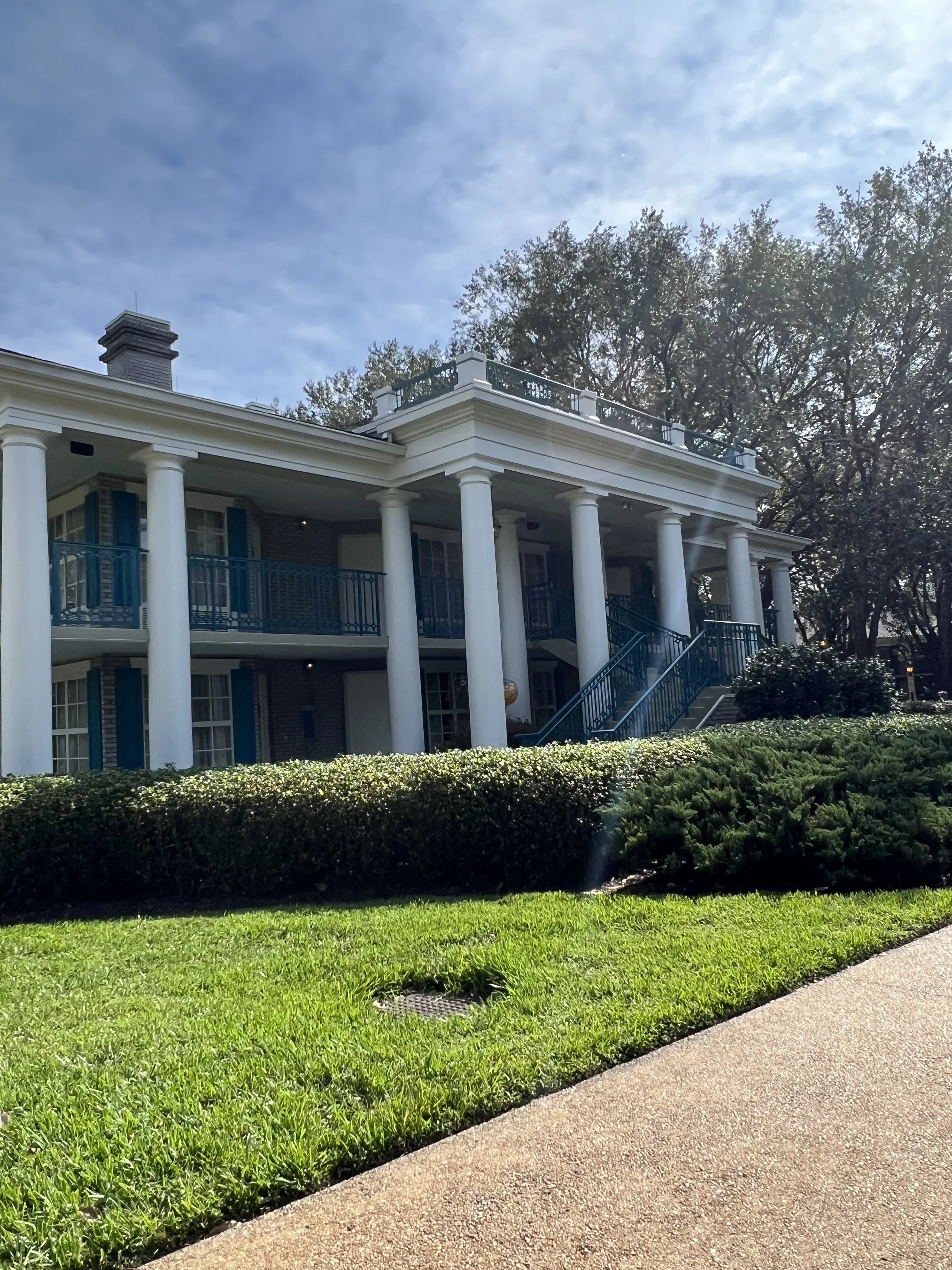 A large white mansion with columns, blue railings, and a staircase, surrounded by greenery and trees under a partly cloudy sky.