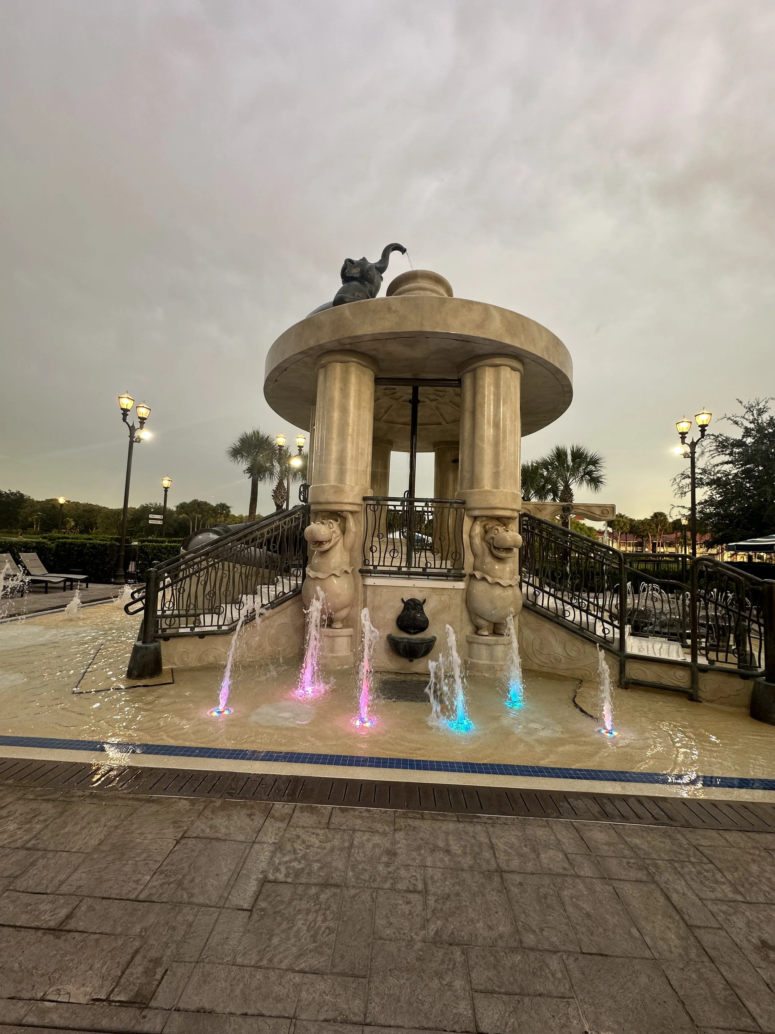 A fountain shaped like a small classical temple with a round roof supported by four columns, adorned with lion heads and a black cat statue on top, and colorful illuminated water jets at the base, in an outdoor plaza with lamp posts, palm trees, and 