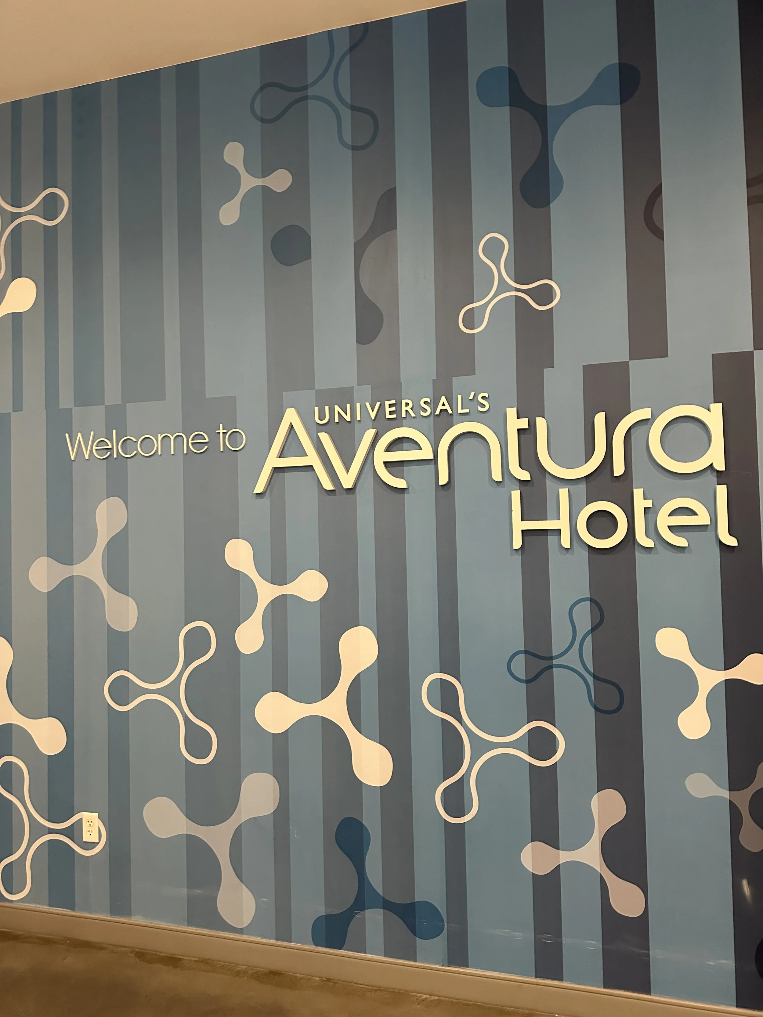 Welcome sign for Universal's Aventura Hotel with a blue background and abstract graphic design.