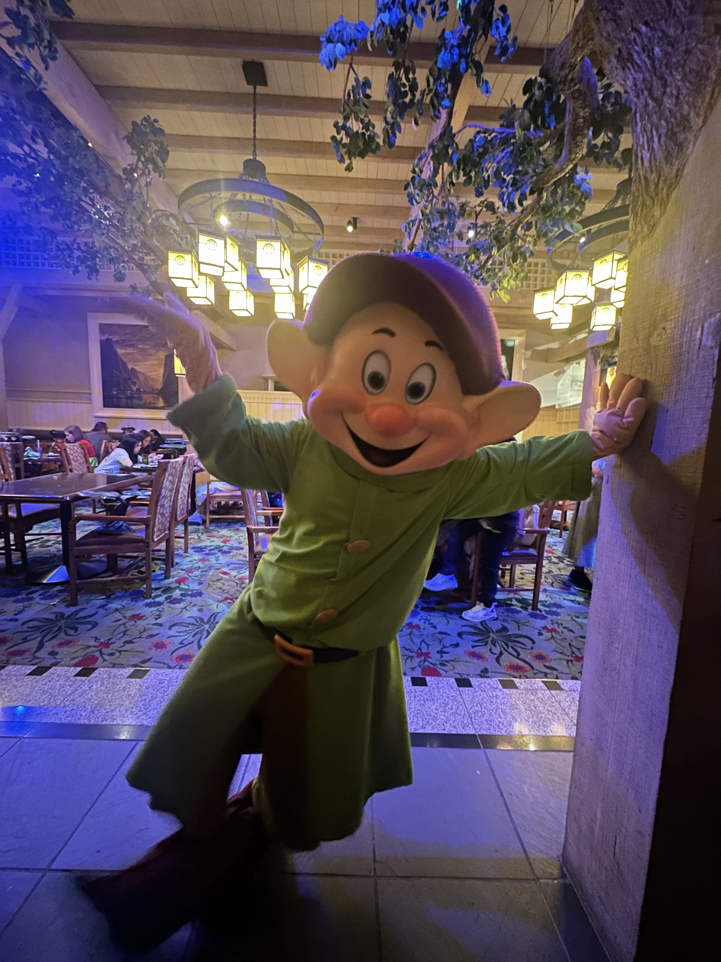 A person in a Lucky the leprechaun mascot costume is posing and smiling at a restaurant or themed venue with tables, multiple people dining, and decorative lighting and paintings in the background.