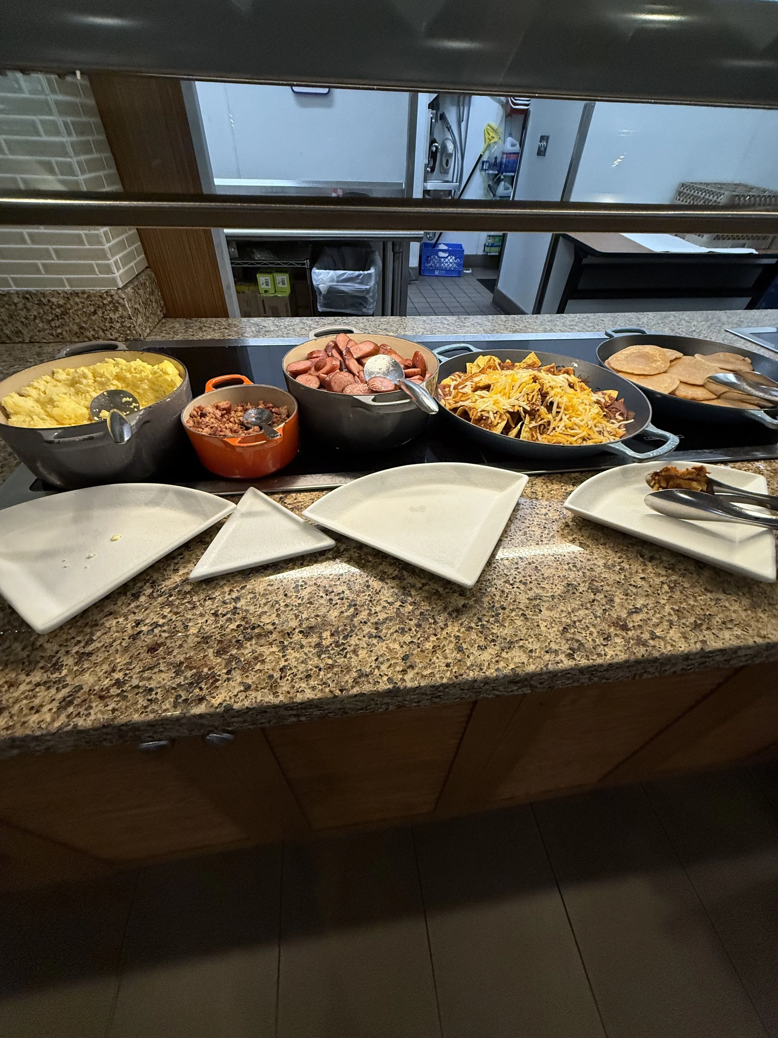 Buffet counter with scrambled eggs, sausages, shredded cheese and meat, ham slices, and empty plates.