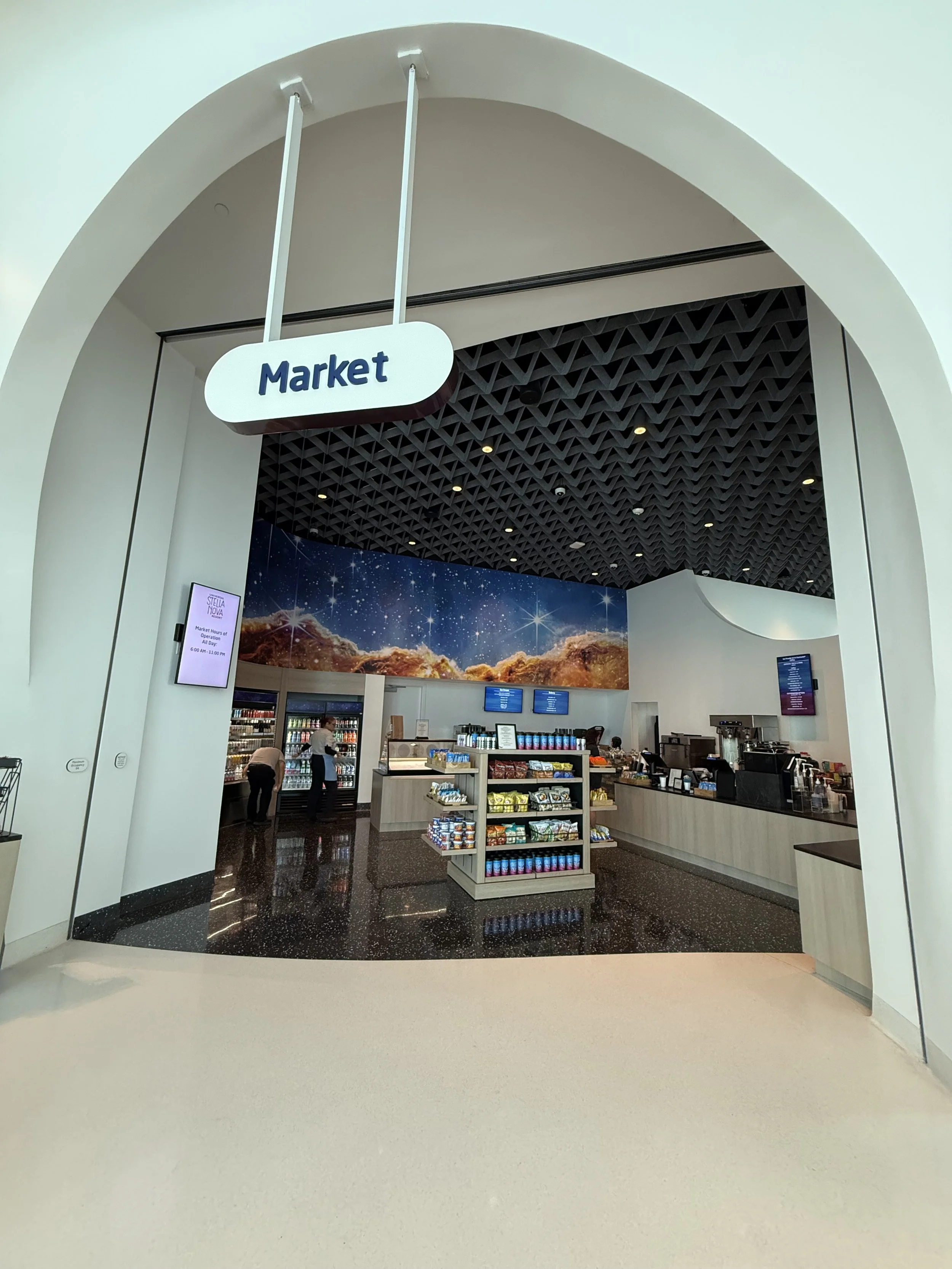 Entrance to a market with a sign overhead, showing a snack and beverage counter inside with digital menus and vending machines.