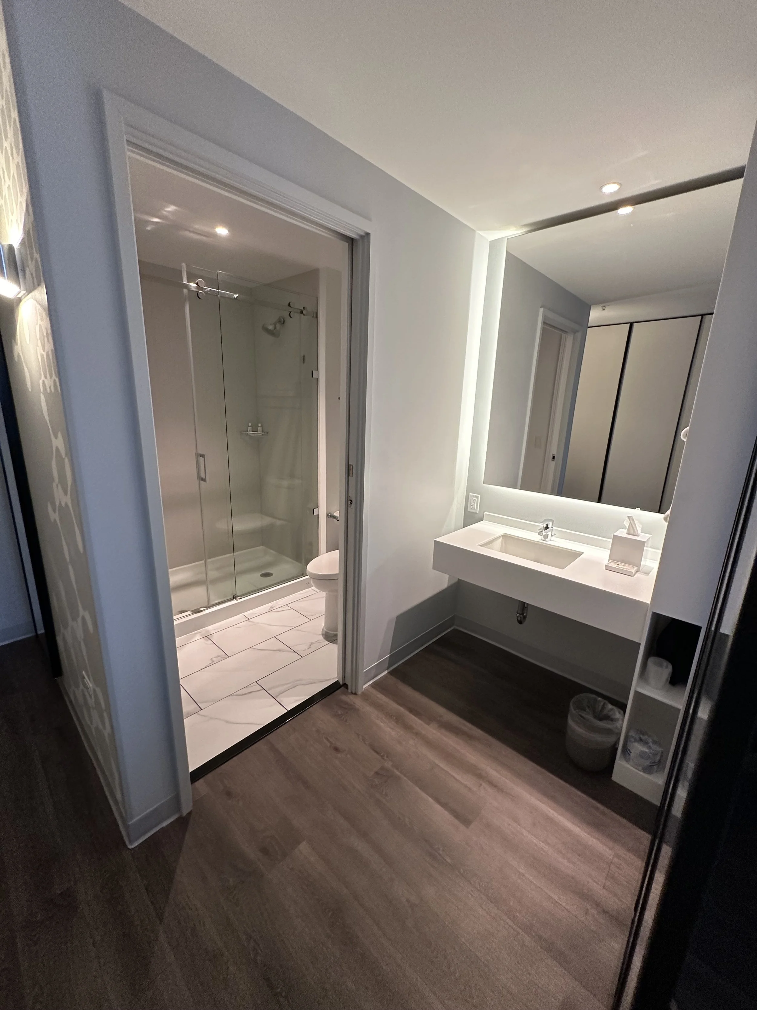 Modern bathroom with wooden flooring, white sink with a mirror, illuminated mirror, tissue box, and a small shelf with toiletries. Visible shower with glass doors and a toilet inside, adjacent to the sink area.