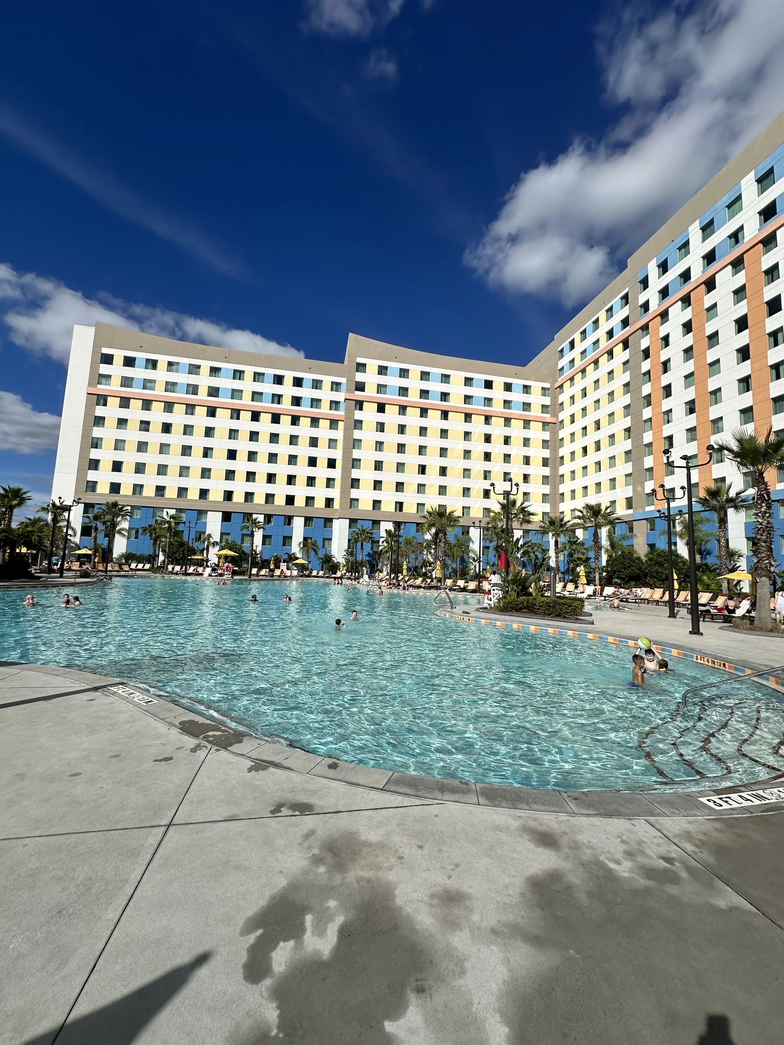 Swimming pool with people enjoying the water, surrounded by lounge chairs, palm trees, and a modern hotel building under a blue sky with clouds.