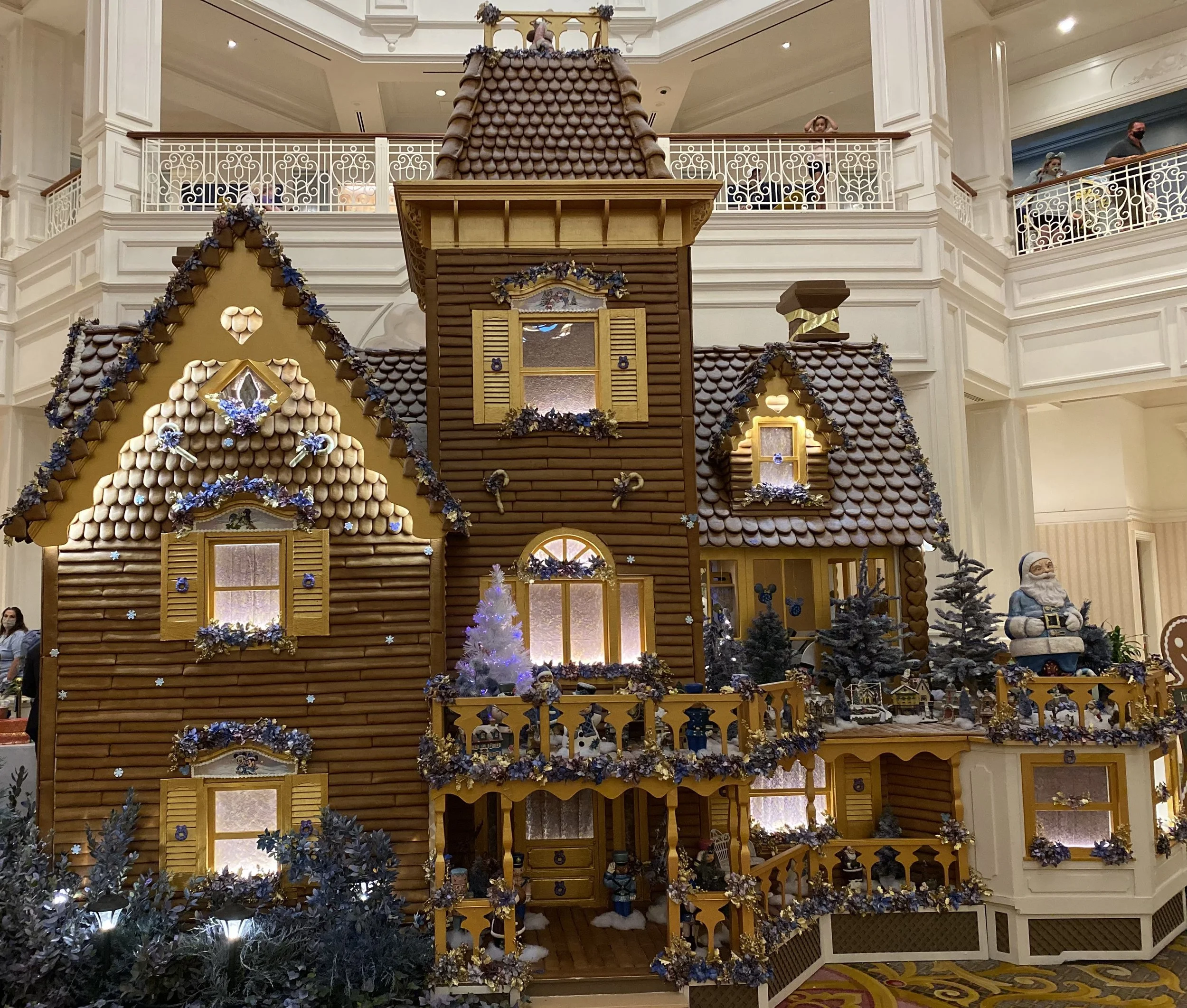 A large gingerbread house display with decorated windows, garlands, and a Santa figurine outside, located in a spacious indoor setting with multiple levels.