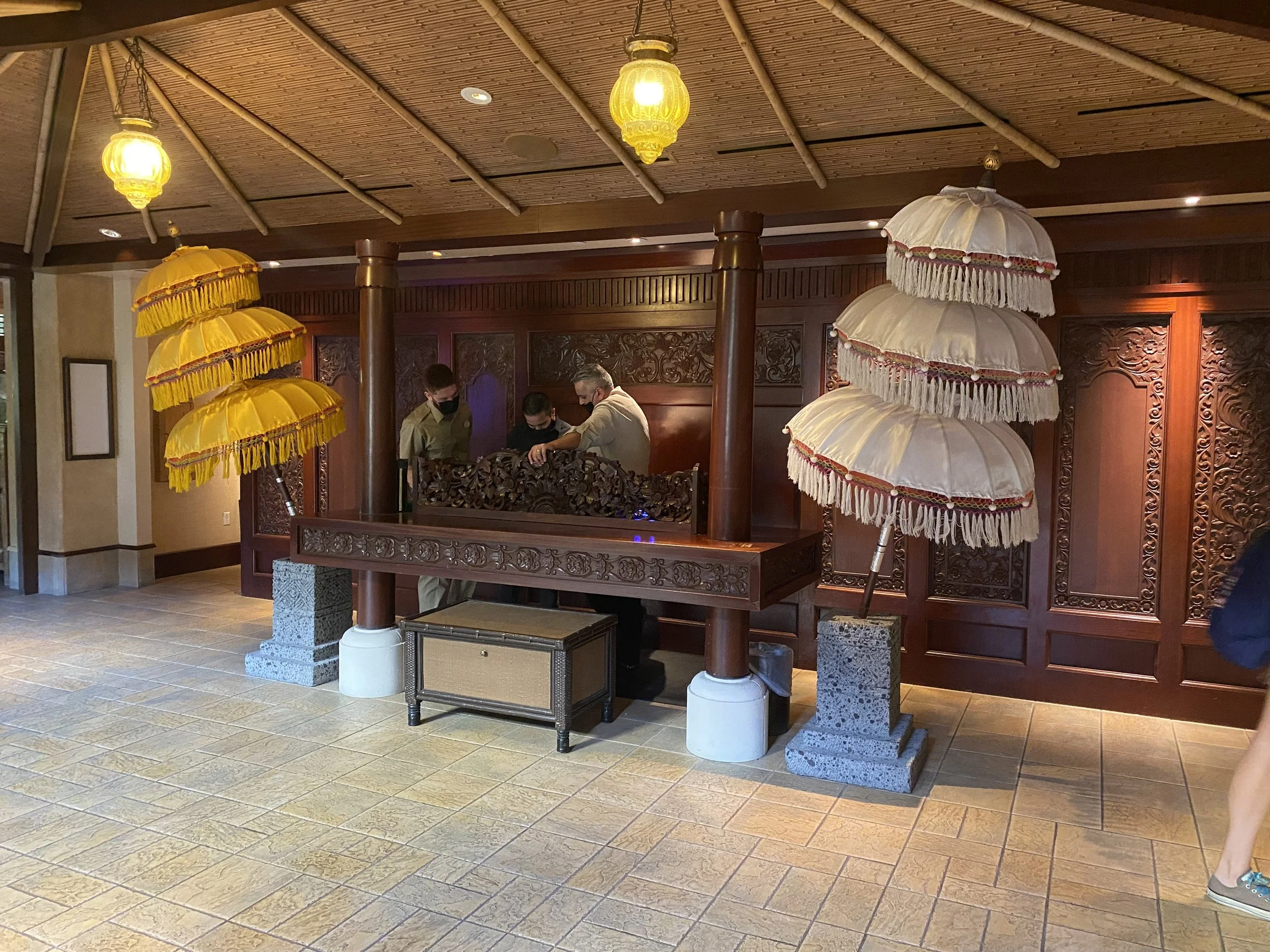 Reception desk with ornate wooden carvings, umbrella stands with traditional decorative umbrellas, and three staff members behind the desk.