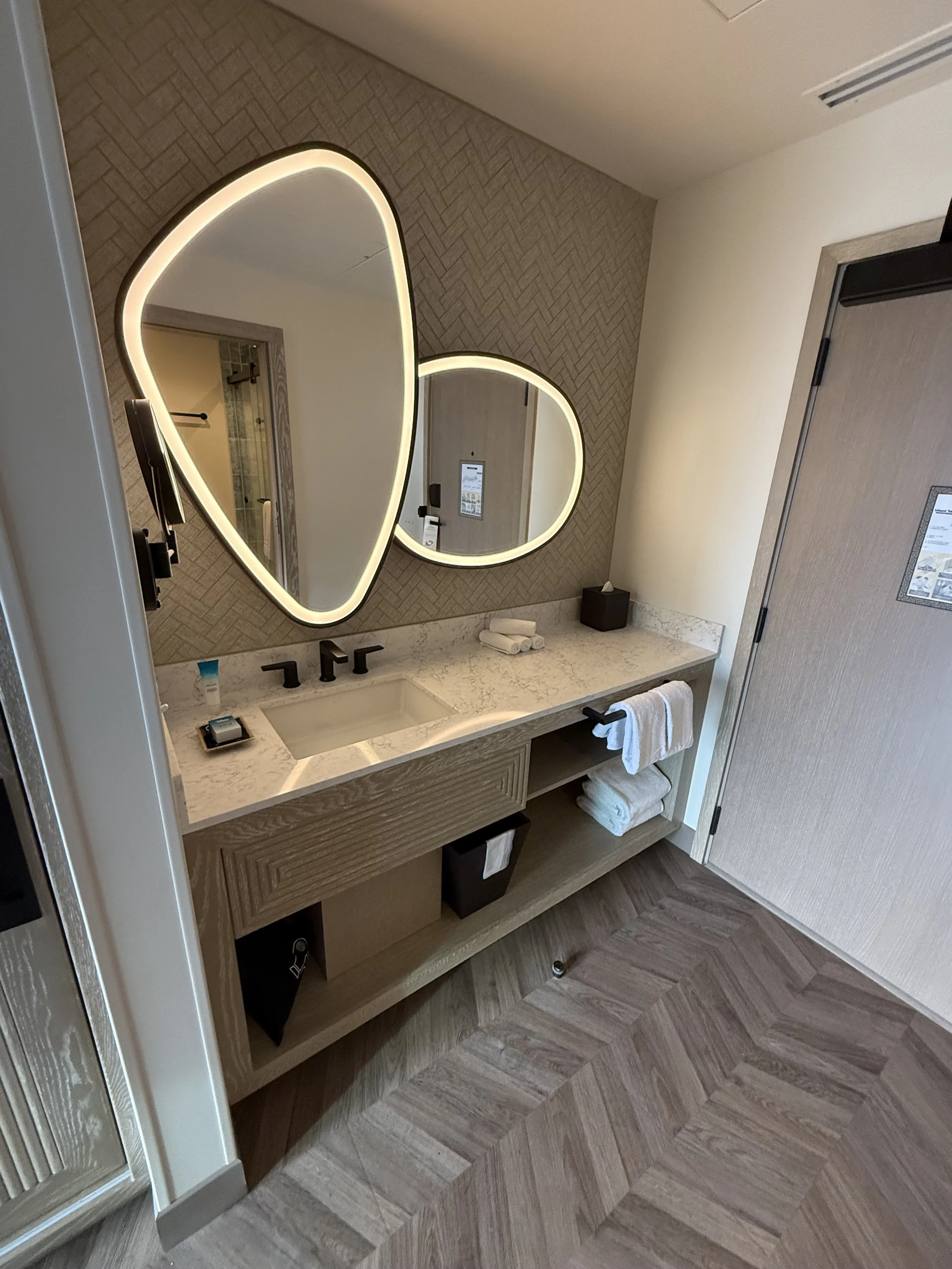 A modern bathroom vanity with a white marble countertop, two black faucets, and two illuminated oval mirrors. The vanity has open shelving with towels and a brown storage bin. The bathroom has wood parquet flooring, a textured accent wall, and light-