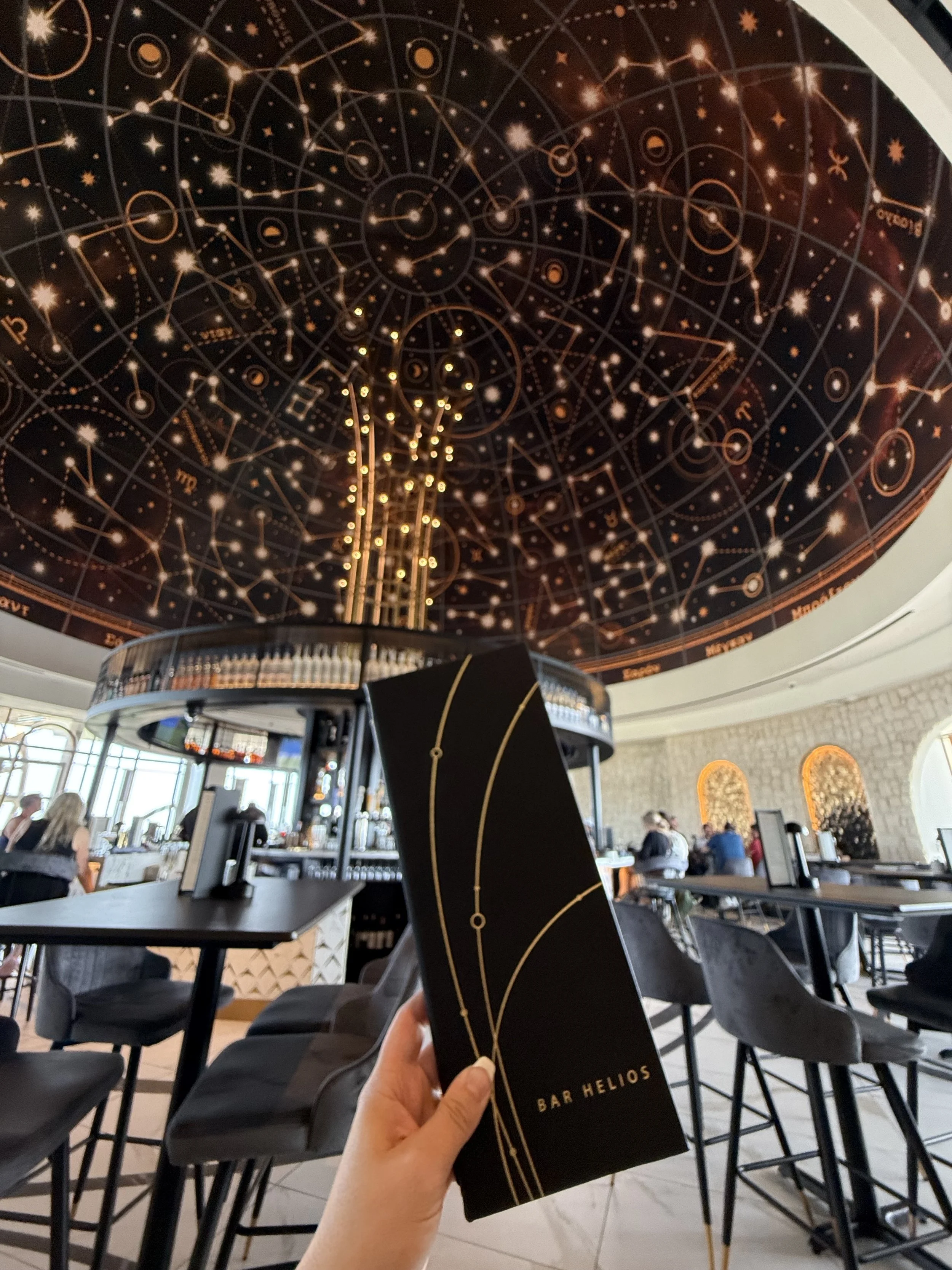 Person holding a black menu with gold lines and "Bar Helios" written on it inside a restaurant with a starry sky or celestial-themed ceiling.