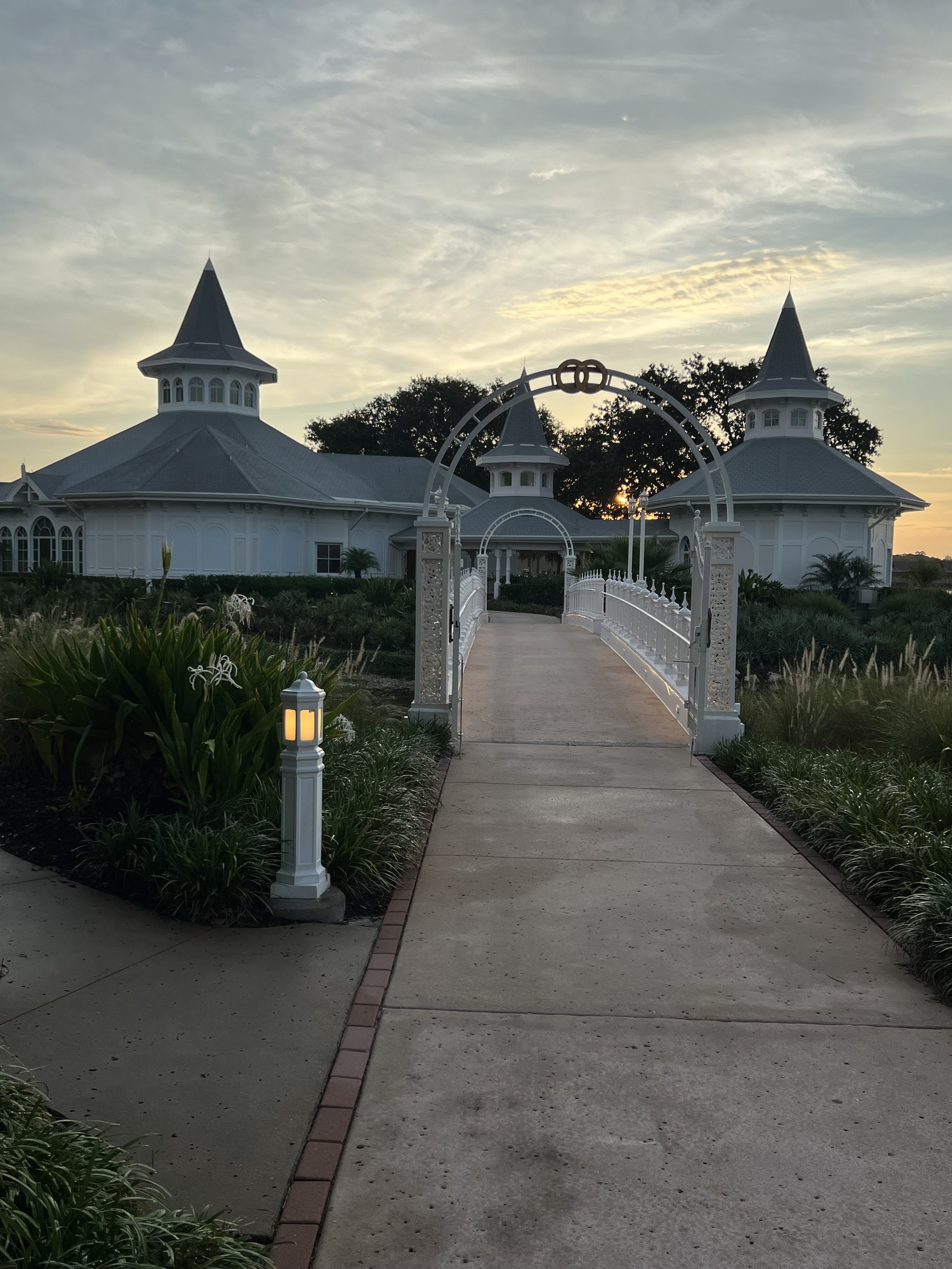 A white decorative bridge with ornate railings leads to a large, white, Victorian-style building with pointed roofs, set against a sunset sky with clouds and trees in the background.