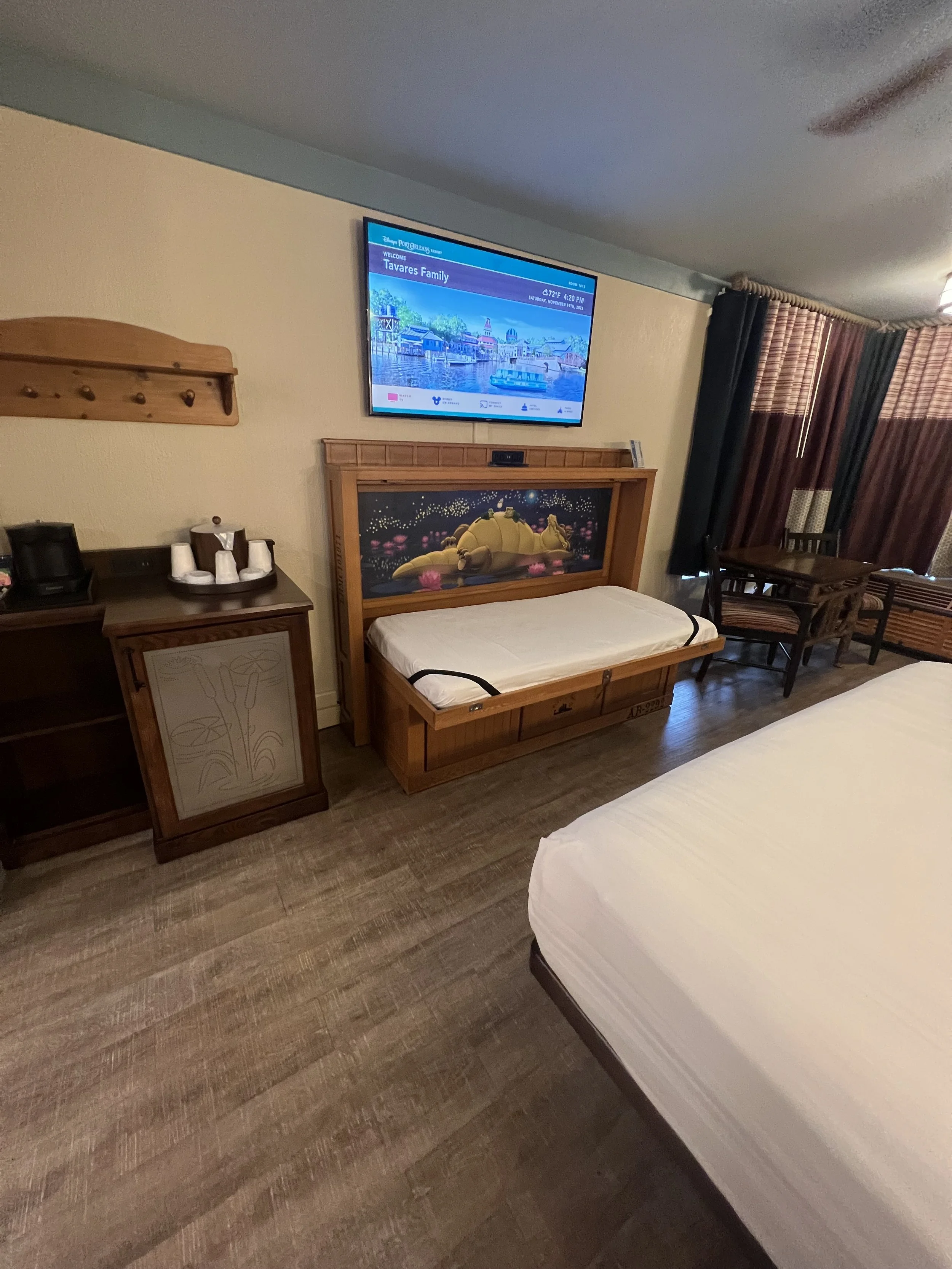 Hotel room with a wall-mounted TV showing a Disney theme park, a wooden bench with a cartoon lion on the headboard, a dresser with cups and a coffee maker, a table with chairs, and curtains covering windows.
