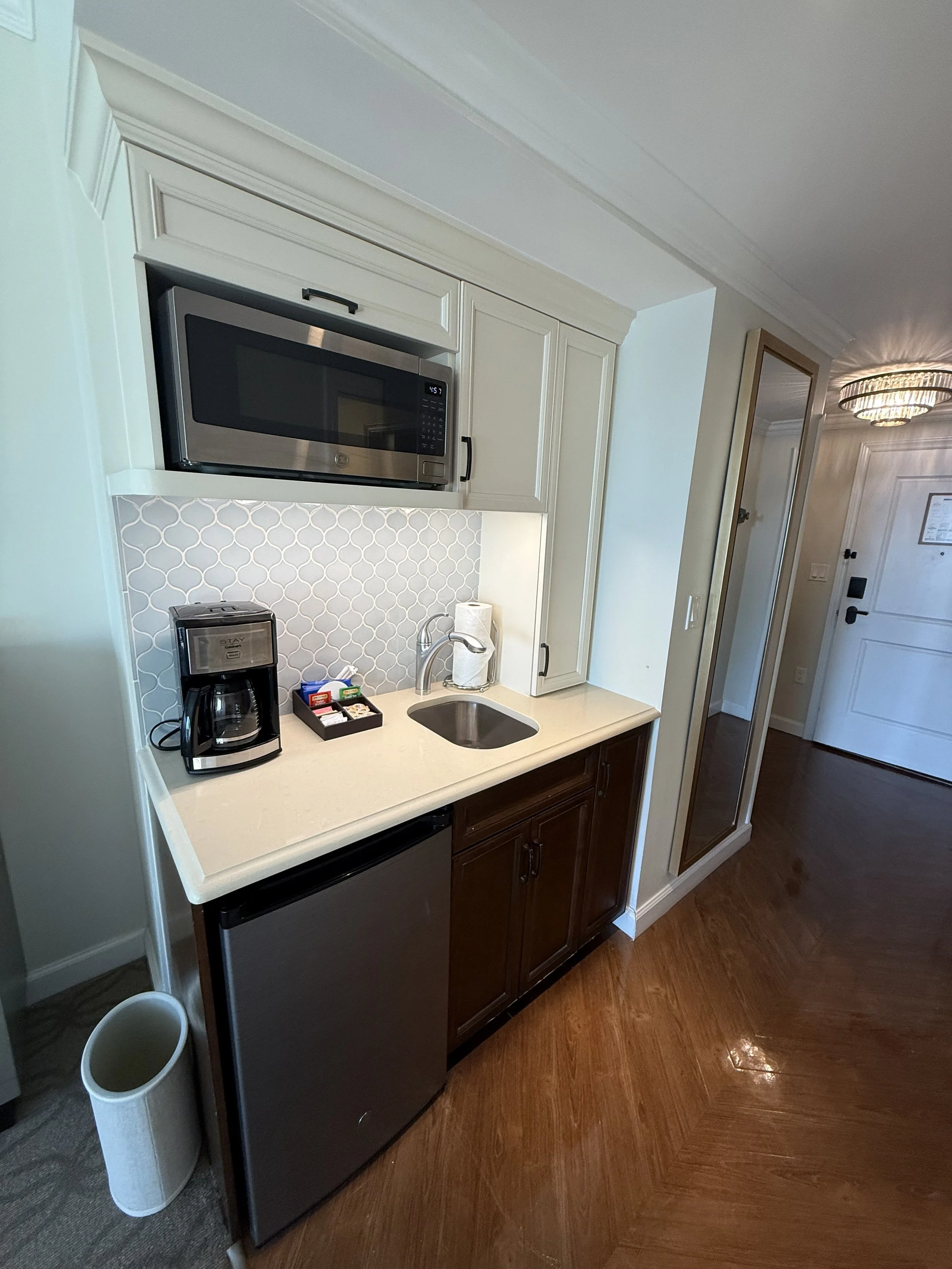 Small kitchenette with a microwave above a countertop, a coffee maker, and a mini refrigerator. There are paper towels next to the sink, and a tray with various coffee pods and accessories. The cabinets are white, and the lower cabinet is dark wood. 