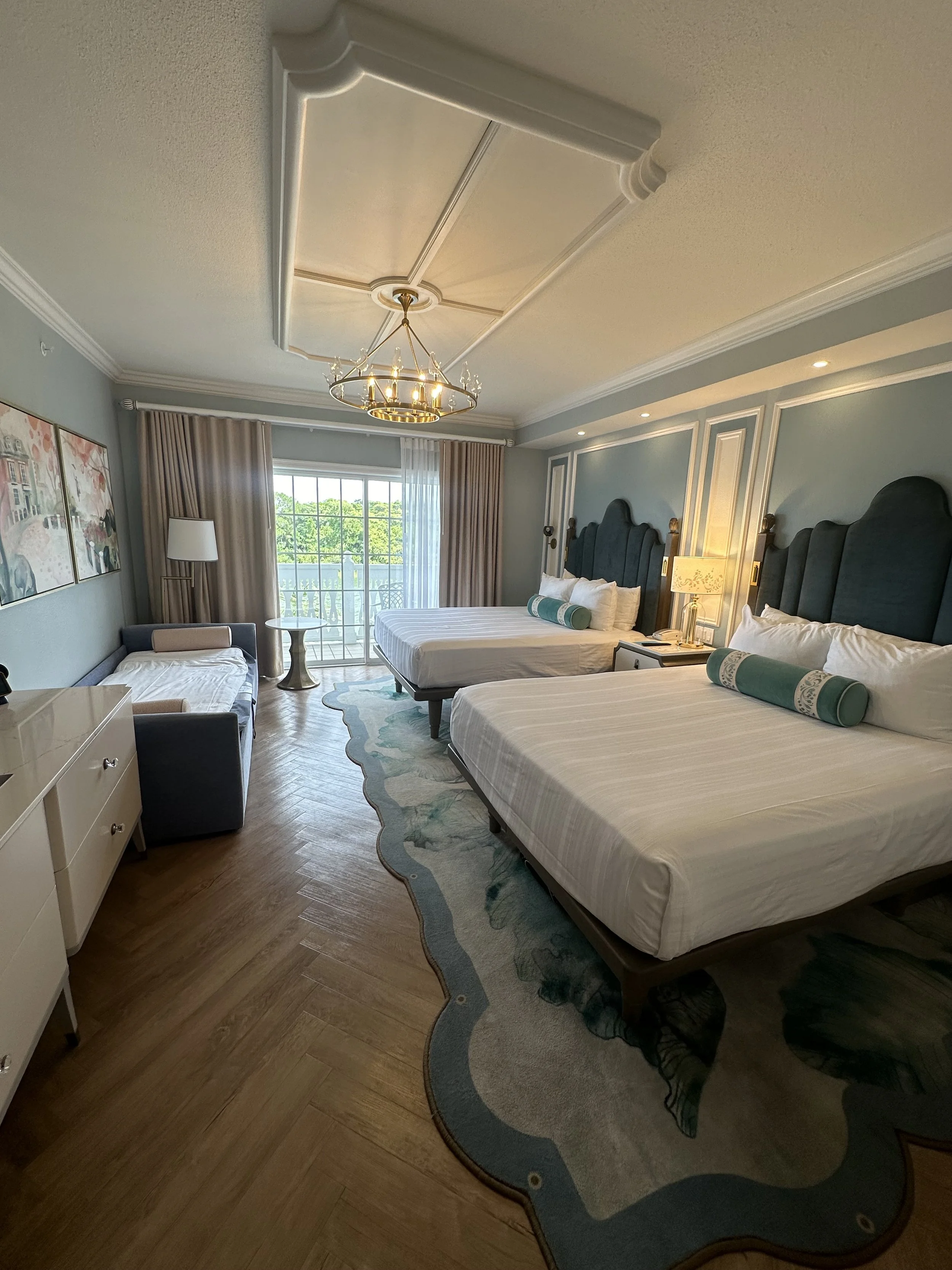 A spacious hotel room with two large beds, a sitting area with a sofa and a lamp, and a balcony with glass doors. The room has light blue walls, wooden flooring, and decorative molding.