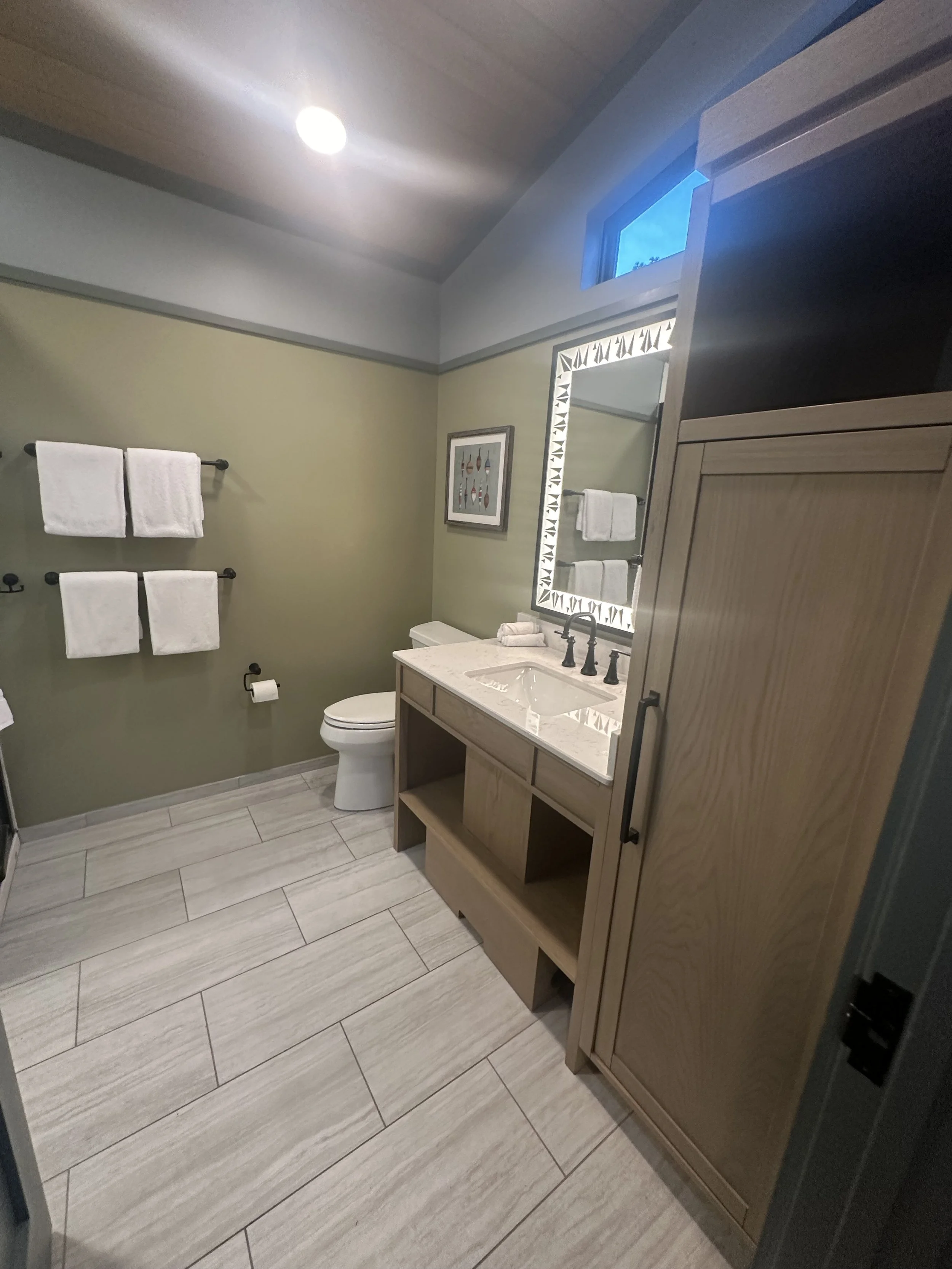 A modern bathroom with beige tiled floor, green wall, towel rack with white towels, toilet, and a beige vanity with a white countertop and sink, seen through an open door.