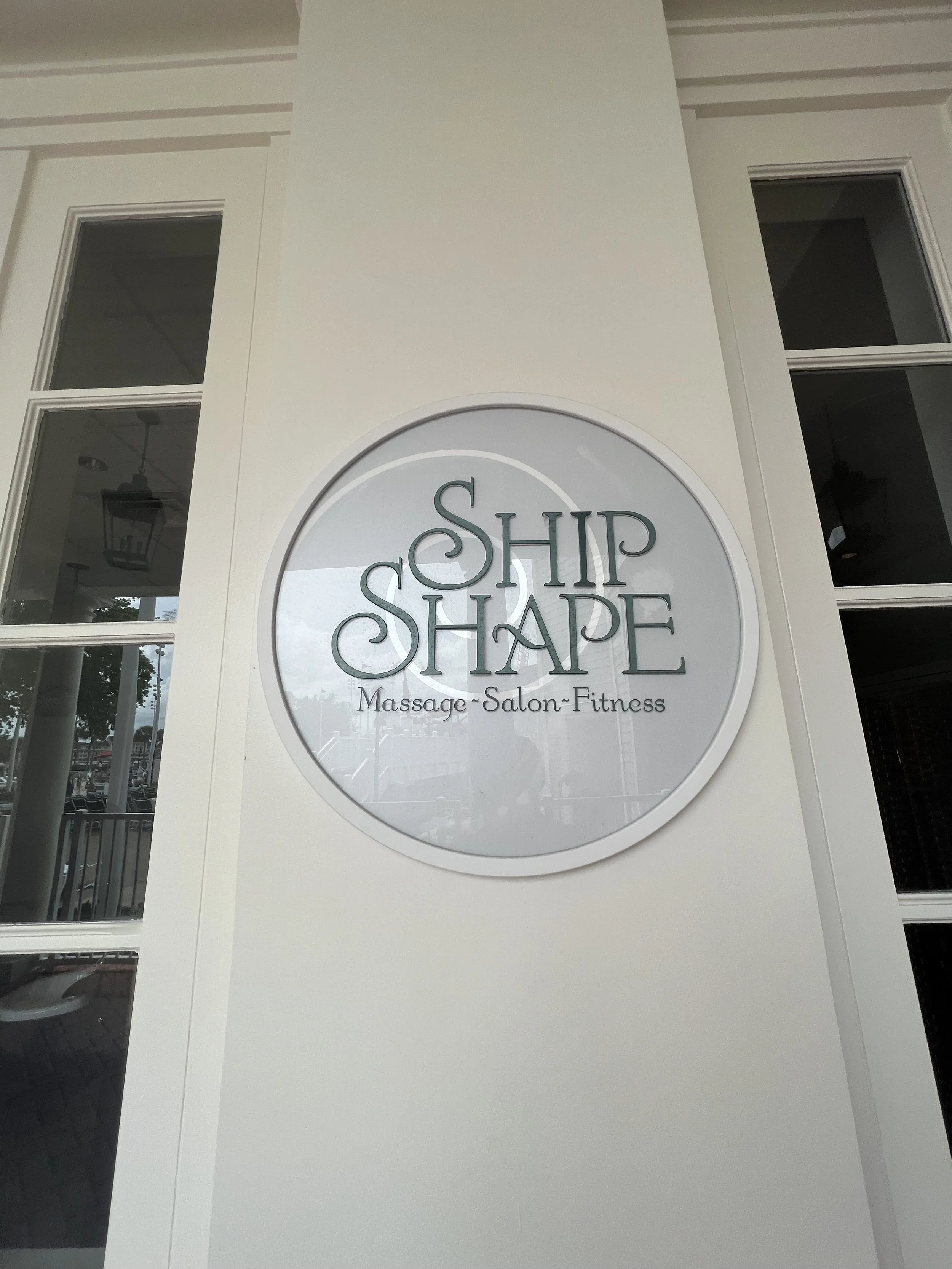 Sign for 'Ship Shape' business offering massage, salon, and fitness services, mounted on a white exterior wall near windows.