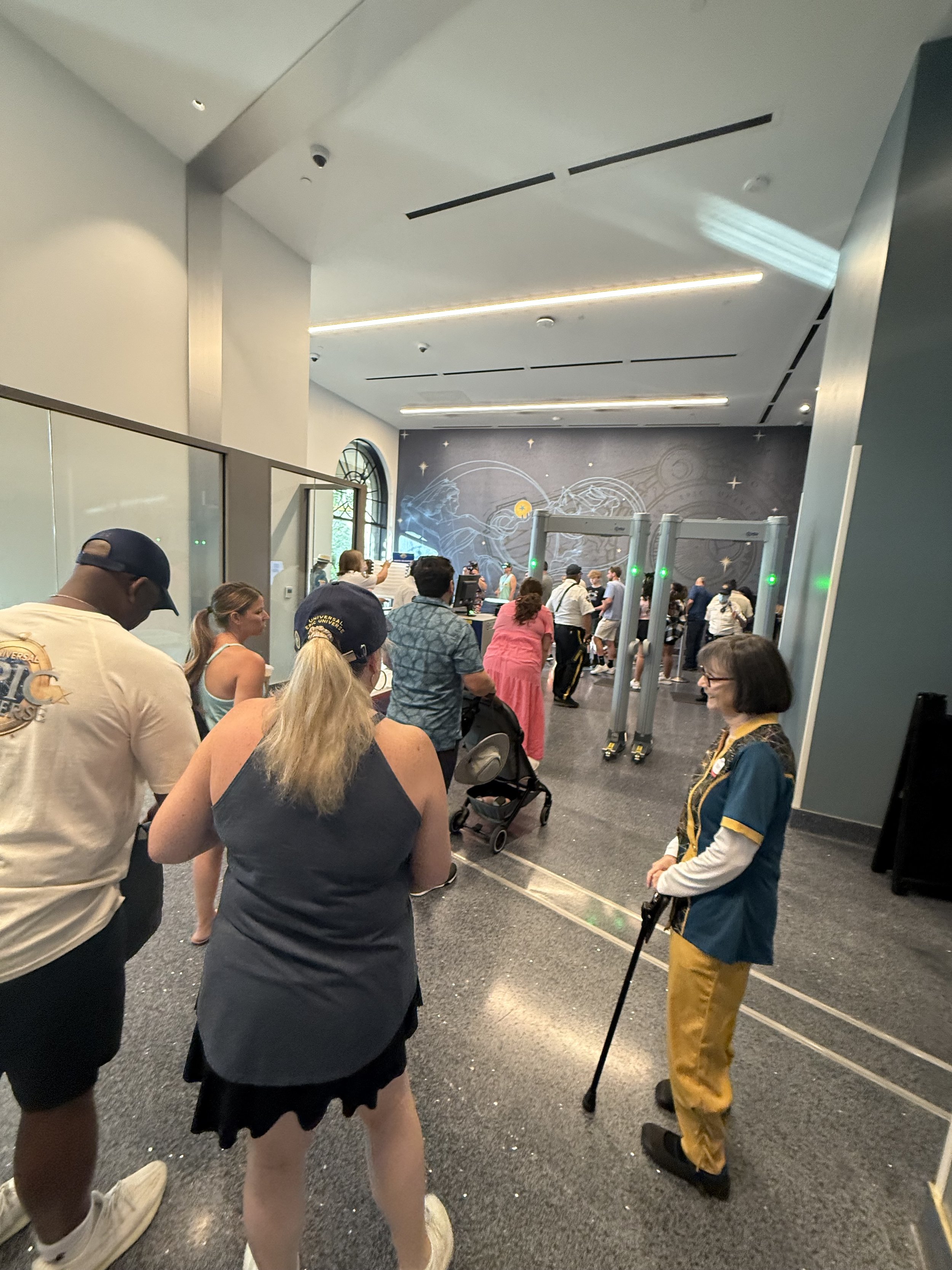 People in line at a theme park entrance with security scanners, with a patterned wall and arched window in the background.