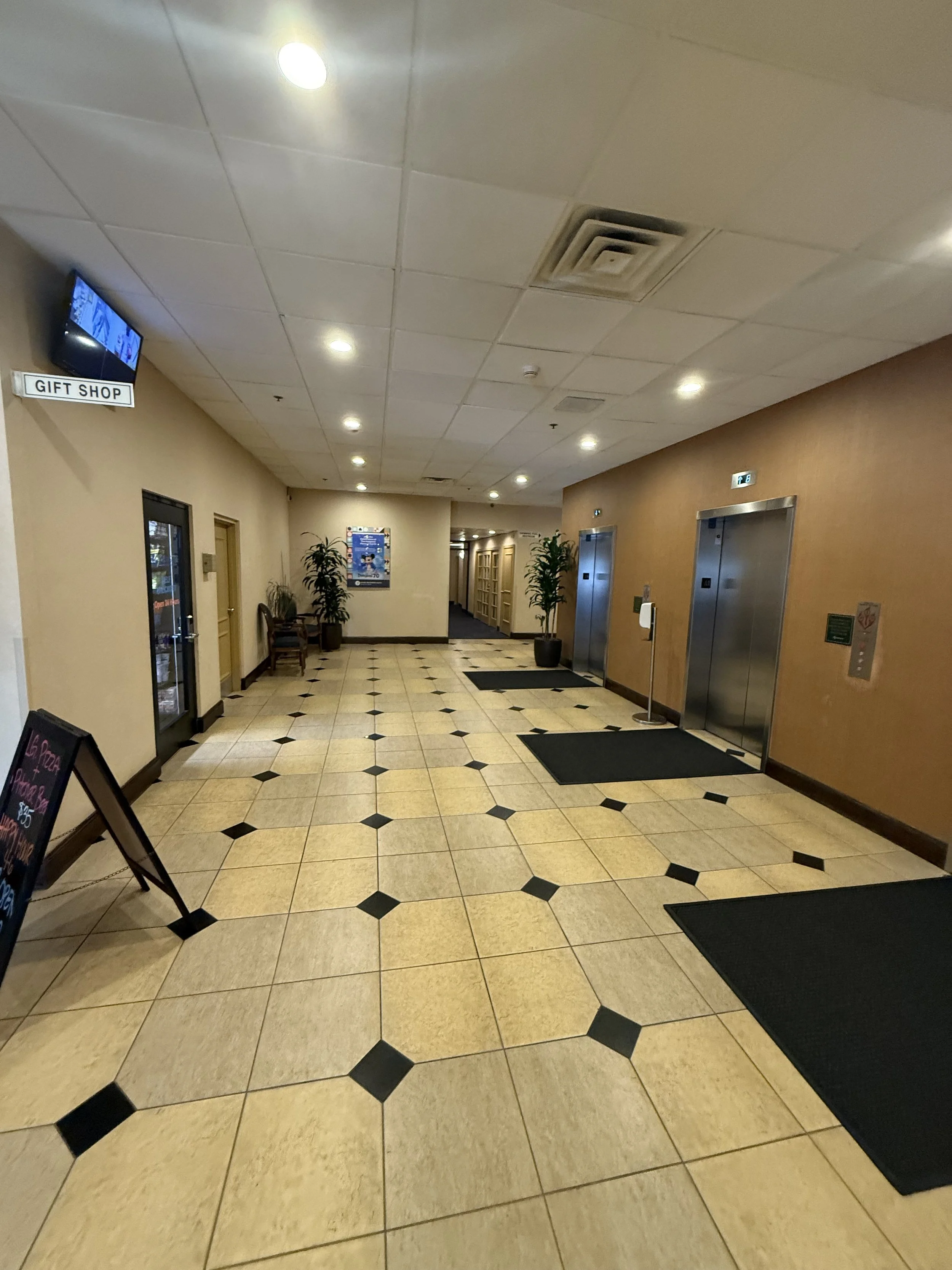 Lobby area with two elevator doors, potted plants, a bench, and a sign indicating a gift shop. The floor has beige and black tile pattern, and there are ceiling lights overhead.