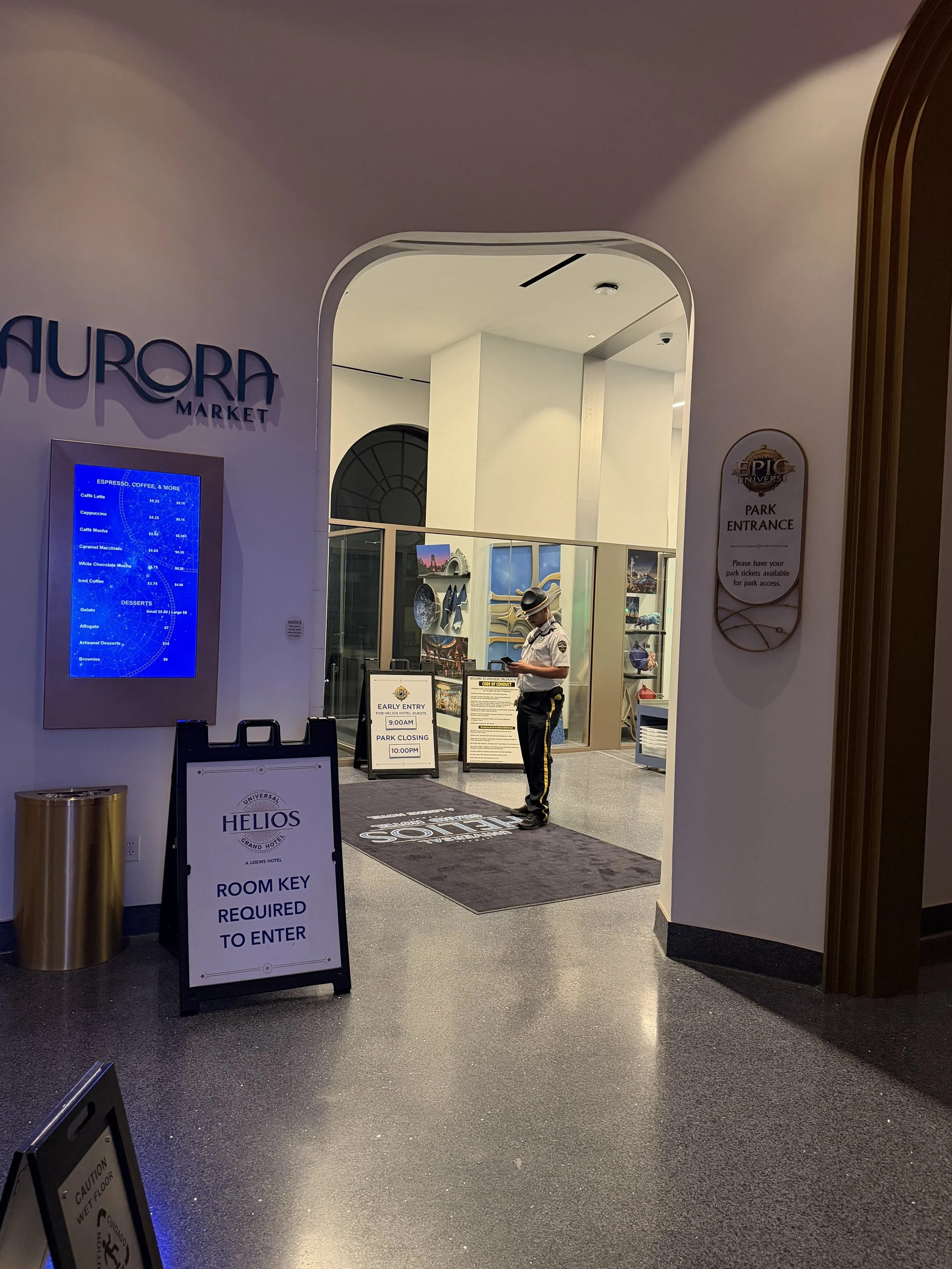 A security guard standing at the entrance of Aurora Market inside a building, with signs about park hours and entrance requirements nearby, and a digital menu board on the wall.