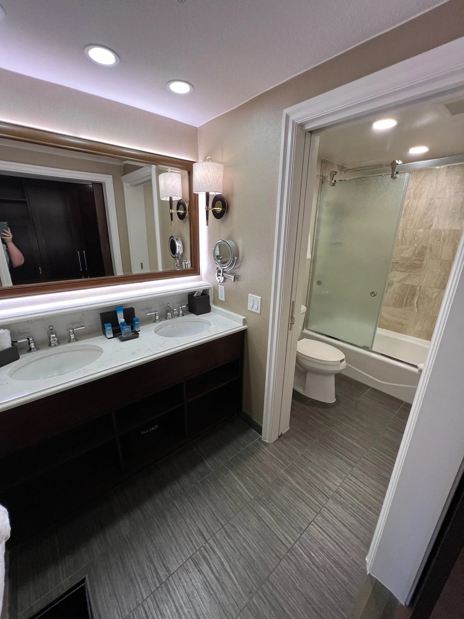 A modern bathroom with a double sink vanity, large mirror, two wall lamps, and a closet with a sliding glass door over a bathtub and toilet.
