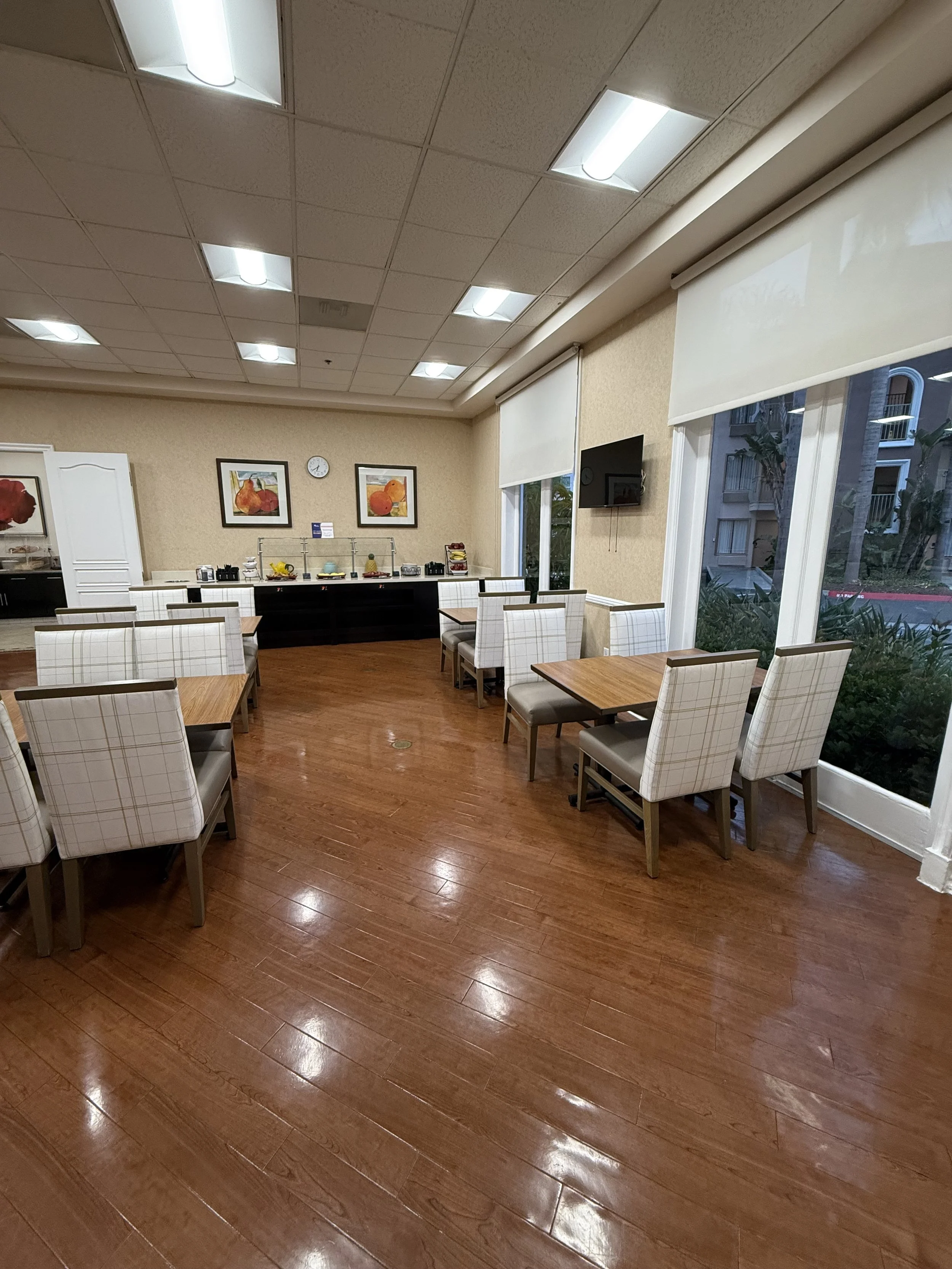Empty dining area with wooden tables, white checkered chairs, large windows, and buffet setup in the background.