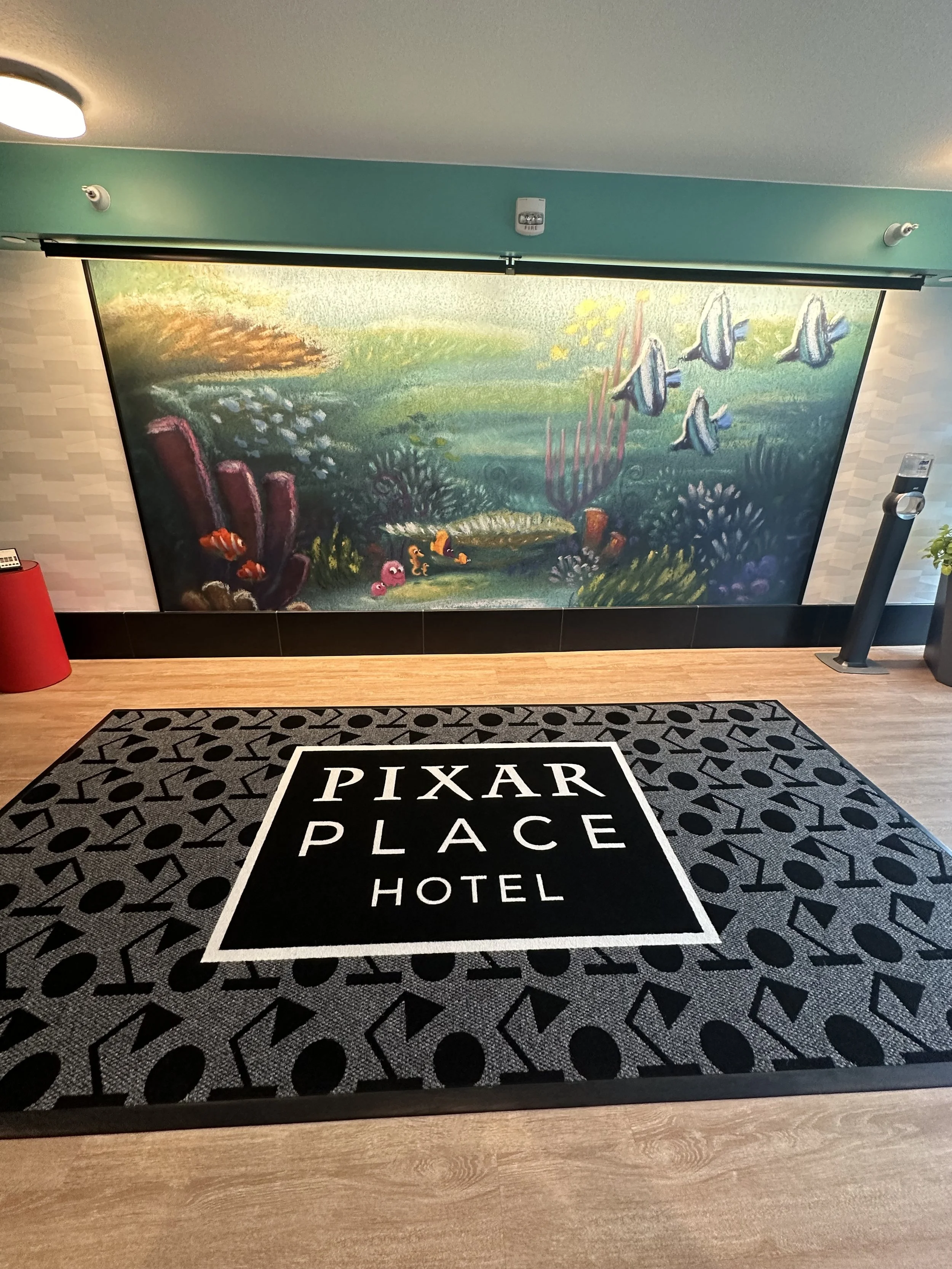 Floor mat with 'Pixar Place Hotel' written on it, in front of an underwater-themed mural featuring colorful coral, fish, and sea creatures, in a hotel lobby.
