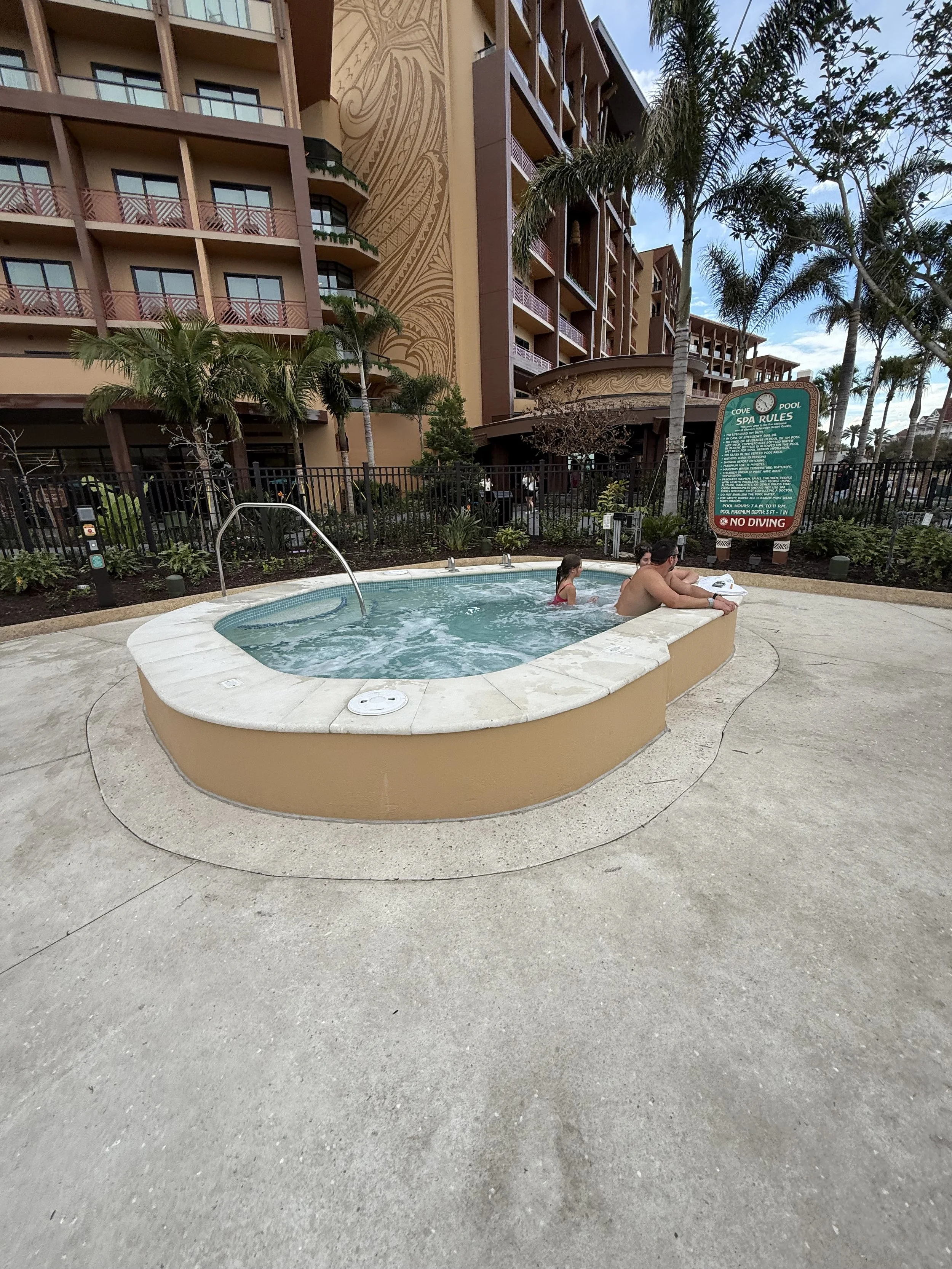 People relaxing in a small outdoor hot tub near a building with palm trees in the background.