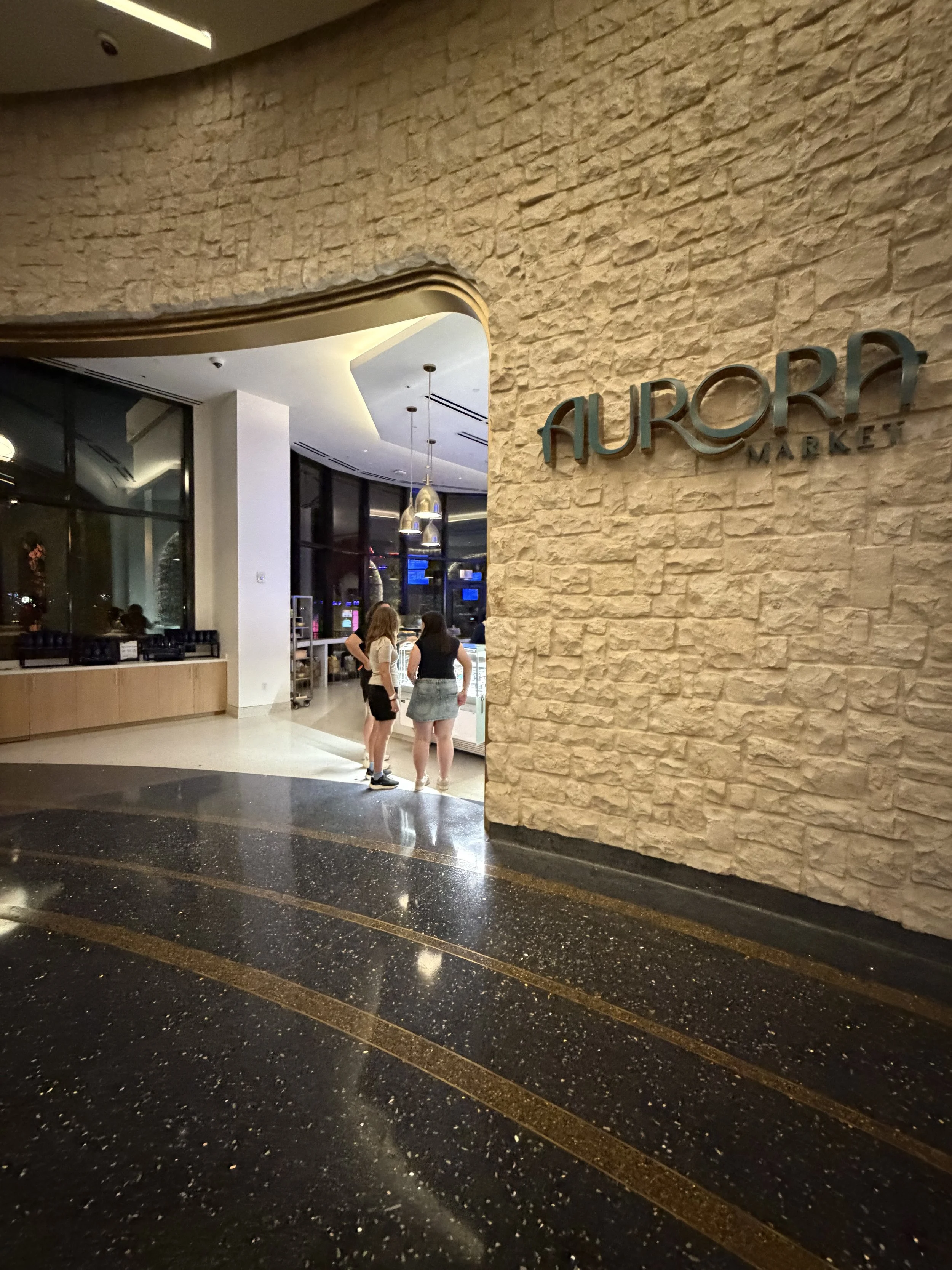 Entrance to Aurora Market with two women standing near the counter, modern interior with stone wall and hanging pendant lights.