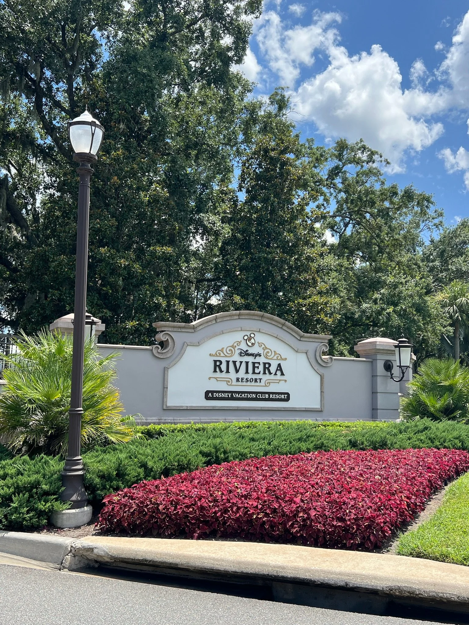 Sign for Disney's Riviera Resort with landscaping and trees in the background under a partly cloudy sky.