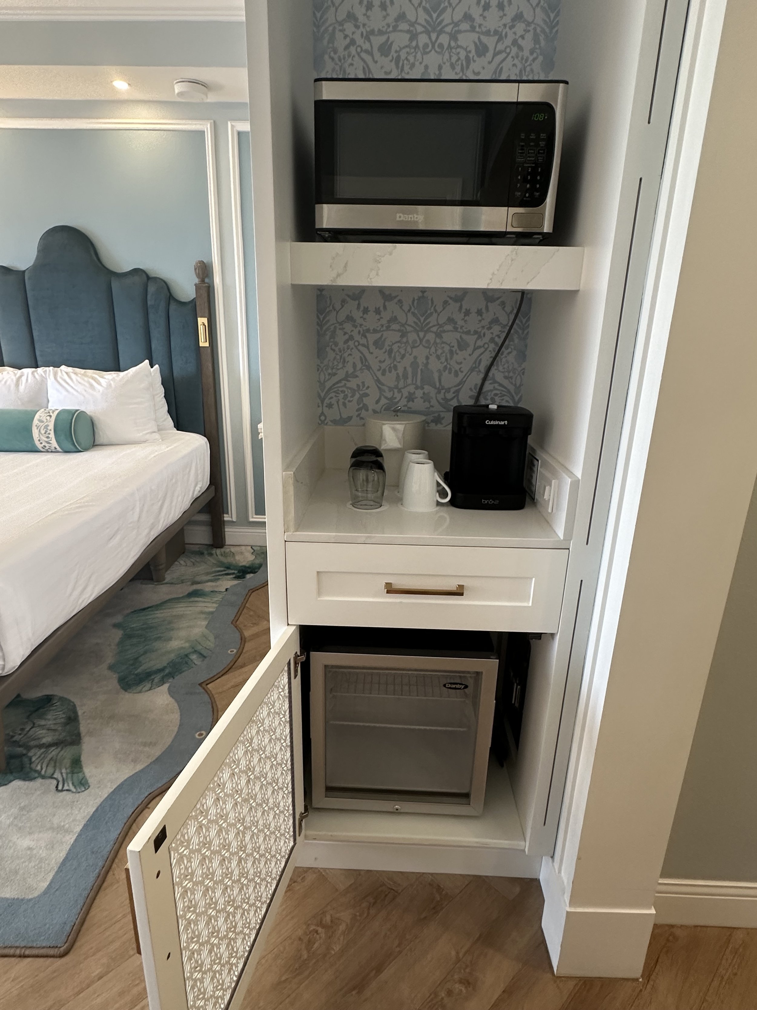 Compact in-room bar area with microwave, mini fridge, coffee maker, water glasses, and cups.