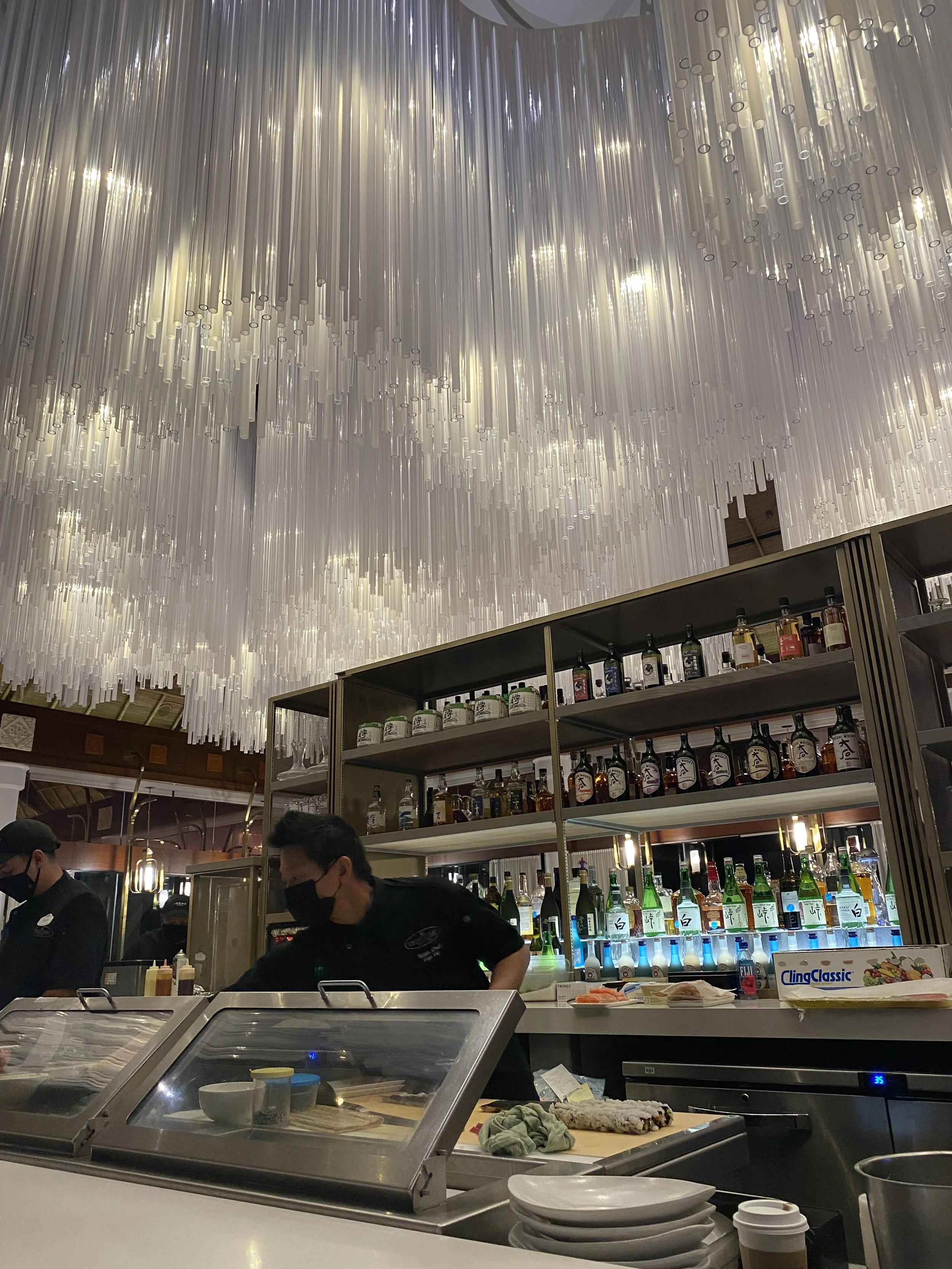 Interior of a restaurant or bar with a decorative ceiling made of hanging white tubes, shelves with various bottles of alcohol, and staff wearing black masks behind the counter.