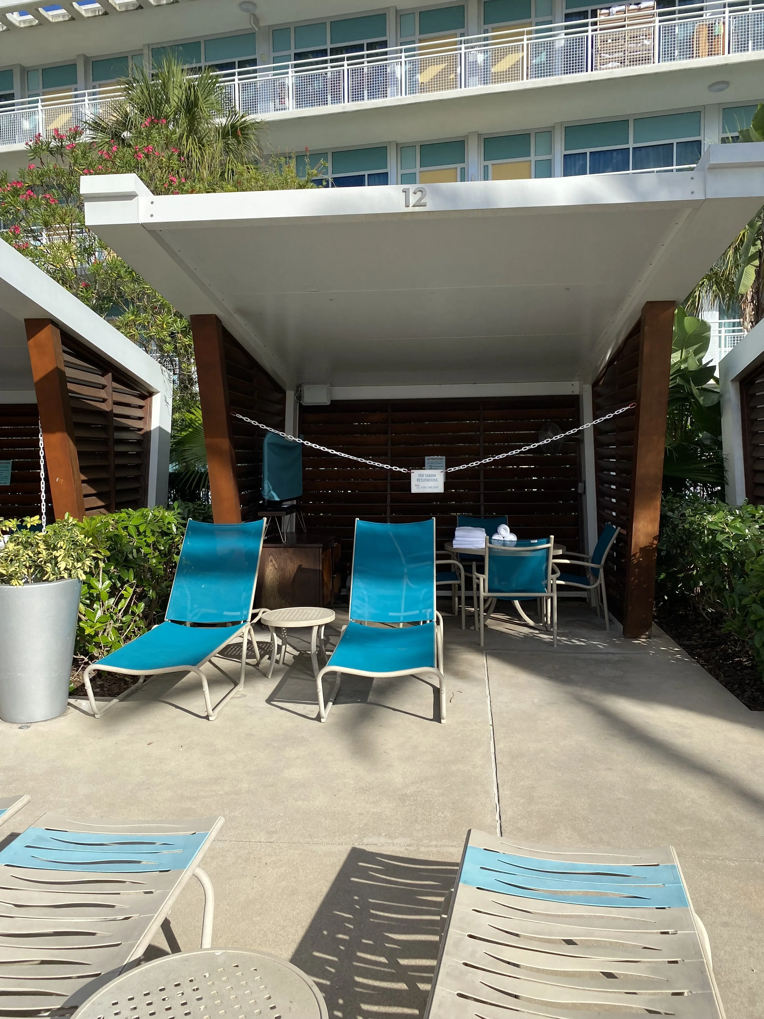 Poolside cabana area with two blue lounge chairs, a small table, a table with towels, and a wooden privacy fence, with a residential building and greenery in the background.