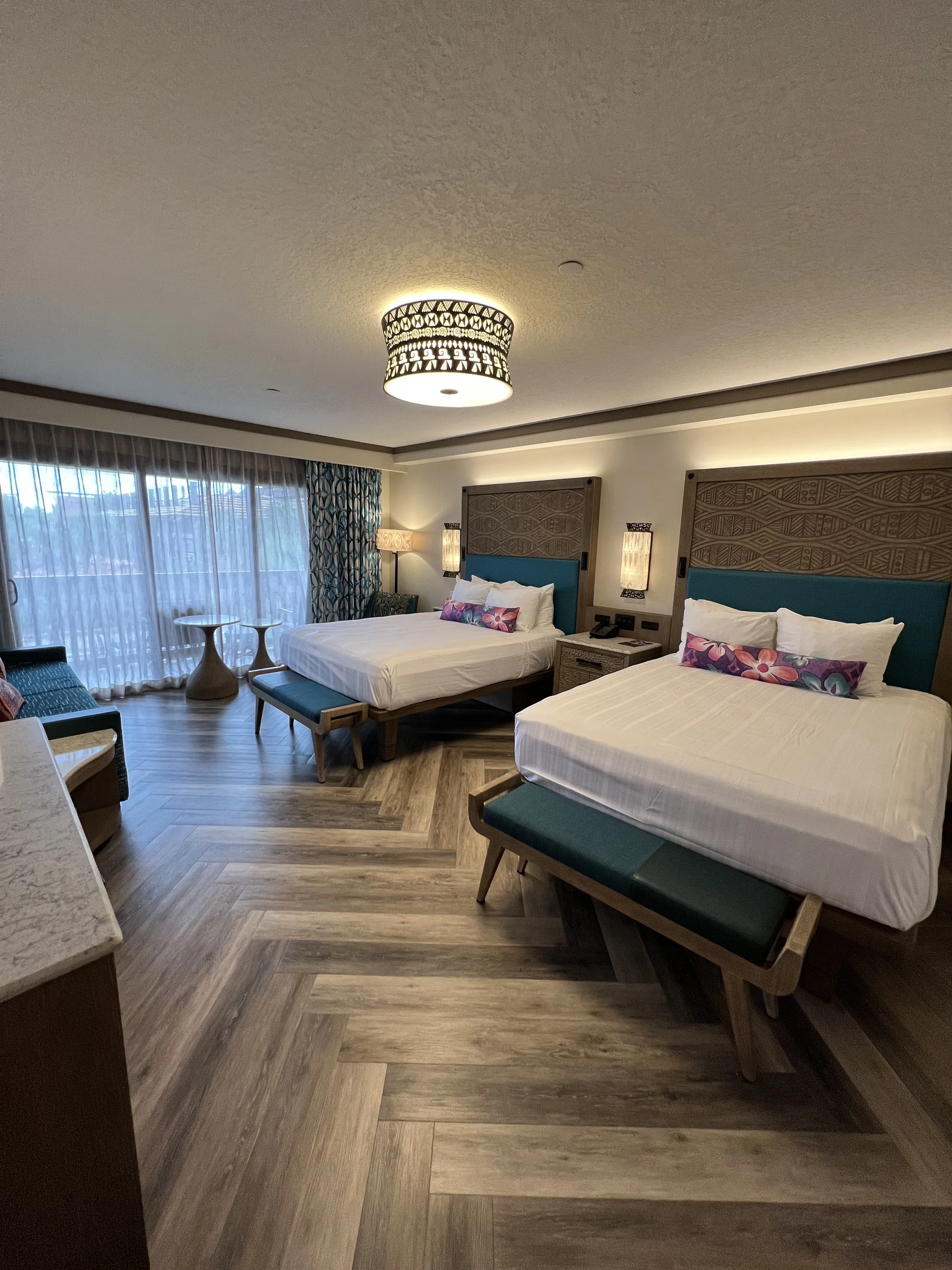 A hotel room with two beds, wooden headboards, and teal accents, facing large windows with sheer curtains, and decorated with contemporary furniture.